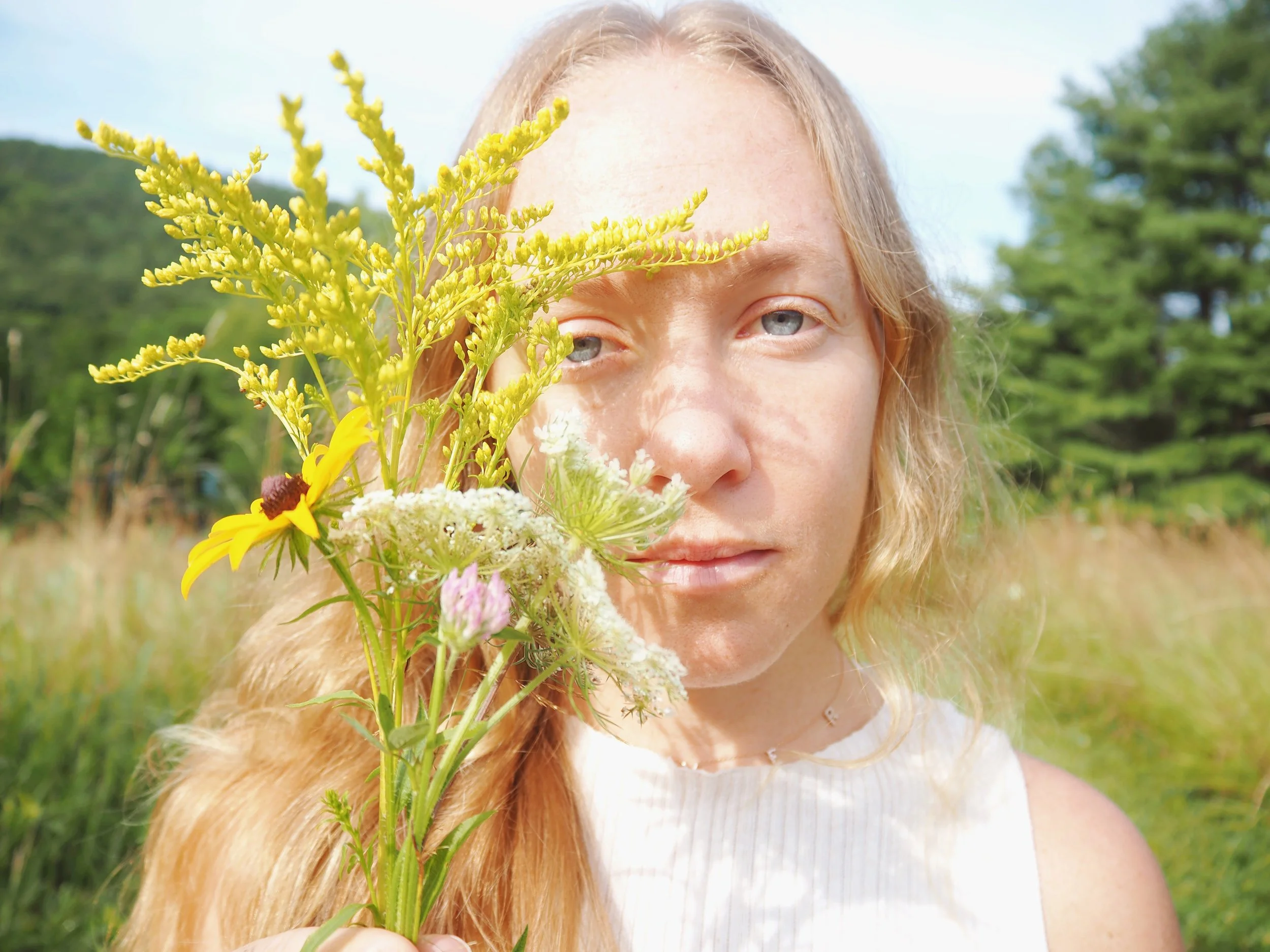 Dru Erin Holistic Brand Coach & Creative Business Doula A woman with light skin and long blonde hair holding a bouquet of wildflowers covering part of her face outdoors on a sunny day.