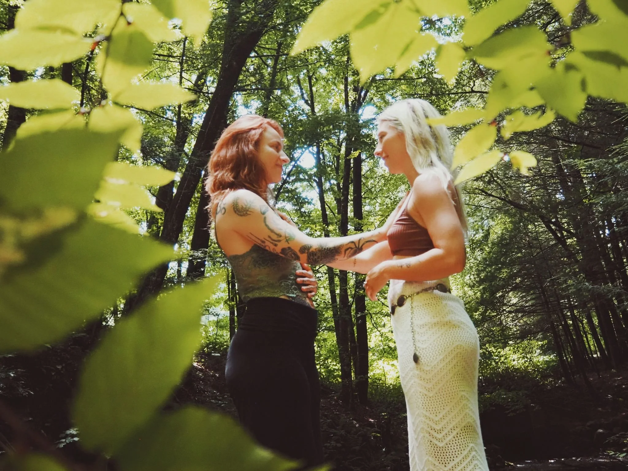 Two women are standing face-to-face in a forest, holding each other's hear and smiling. Relational support and trust between women, reflecting intuitive business mentorship and high-touch consulting for female founders