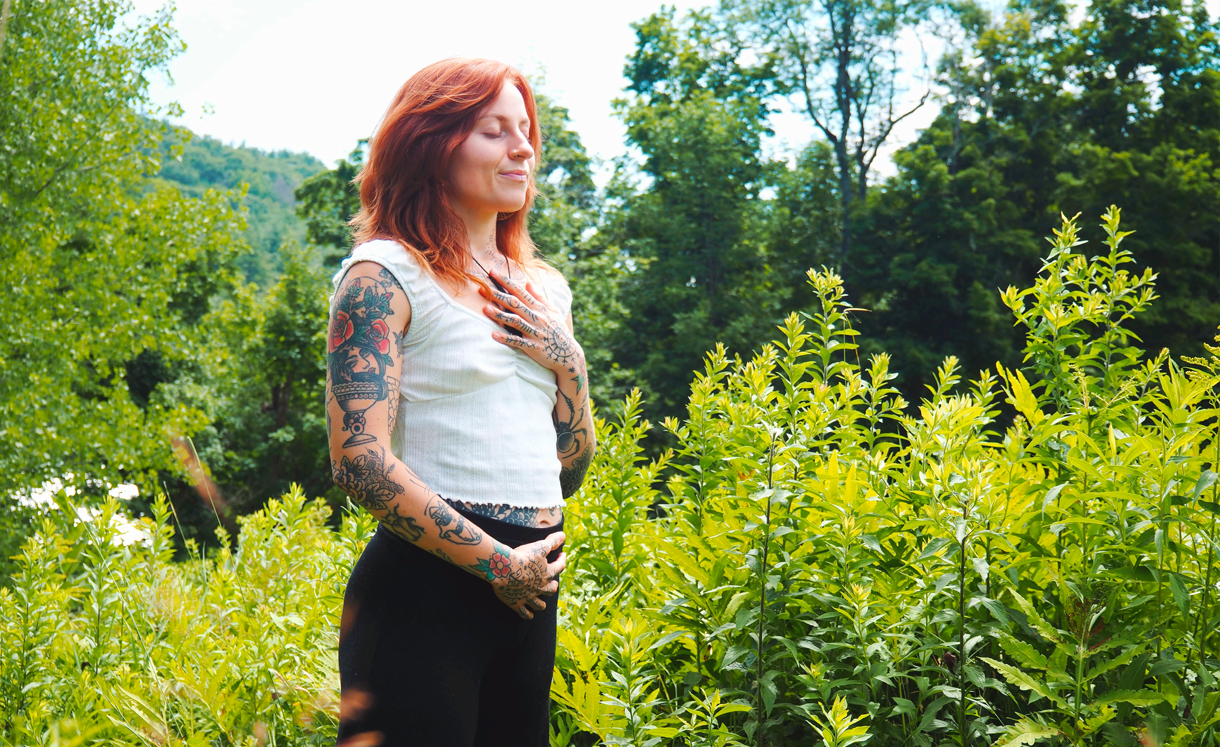 A woman standing outdoors with her hand on her chest, eyes closed, surrounded by lush green trees and bushes. Grounding and nervous-system regulation for women entrepreneurs rebuilding businesses that support their life and capacity