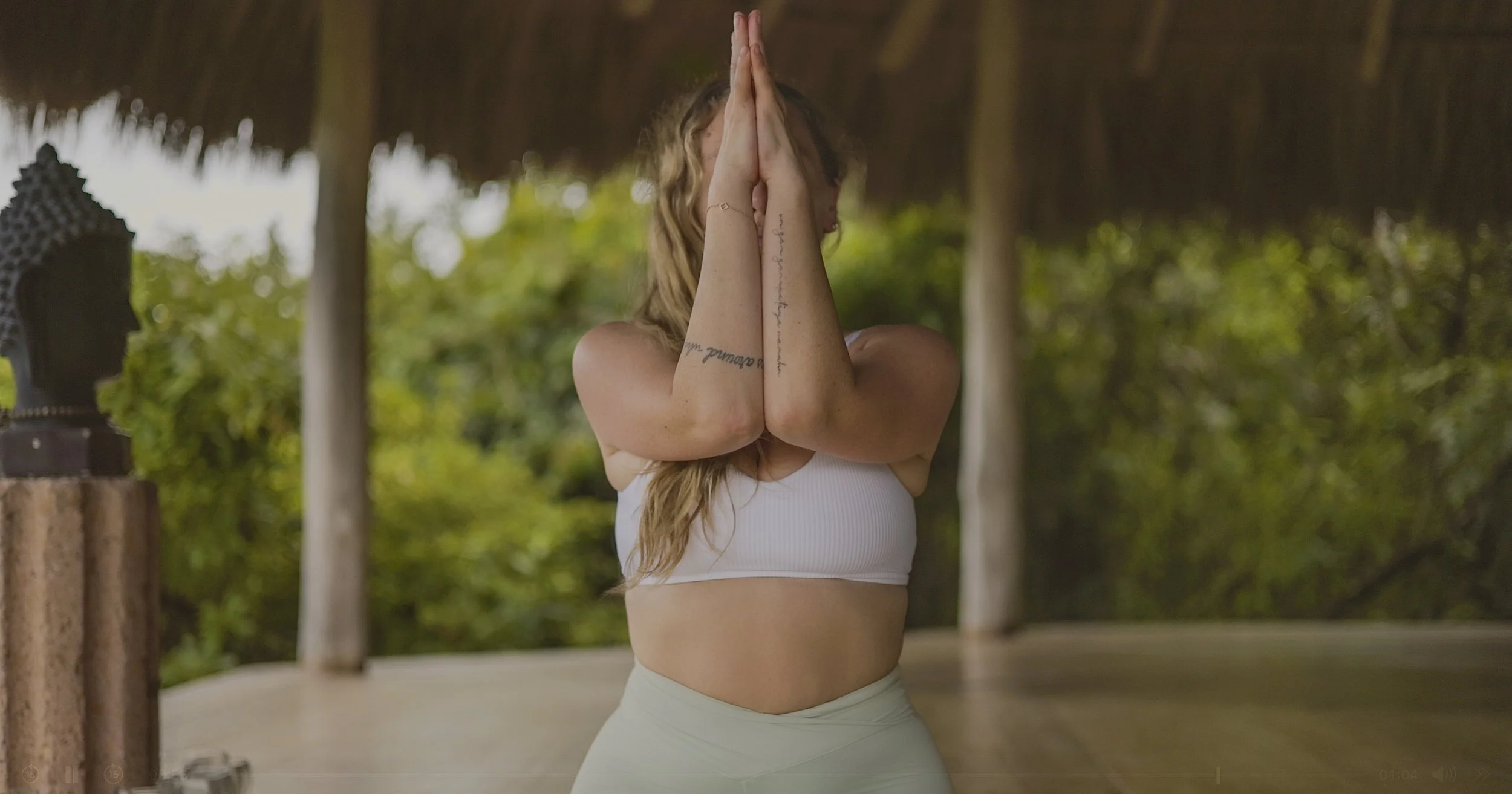 Dru Erin Holistic Brand Coach & Creative Business Doula performing yoga outdoors, standing with hands pressed together in prayer pose, wearing a white crop top and light-colored leggings, with a thatched roof and green foliage in the background.