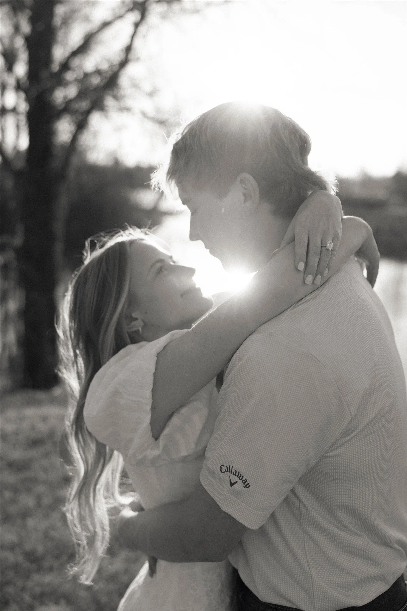 A bride and groom dancing outdoors on a grassy field under a large tree, with sunlight filtering through the leaves.