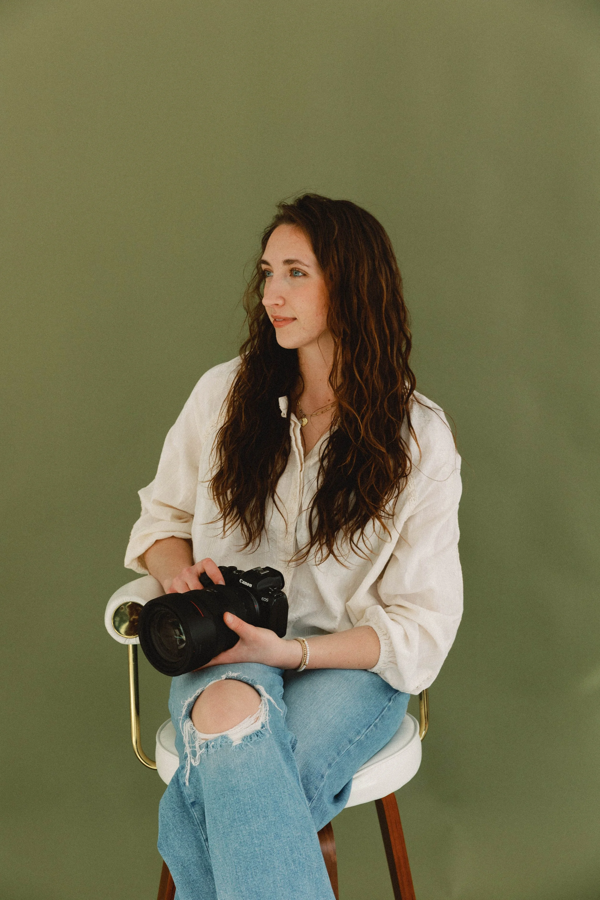 A woman with long, wavy brown hair and blue eyes sitting on a white chair with a gold armrest, holding a professional camera, looking to her left, wearing a white oversized shirt with rolled-up sleeves and ripped jeans against a plain light olive green background.