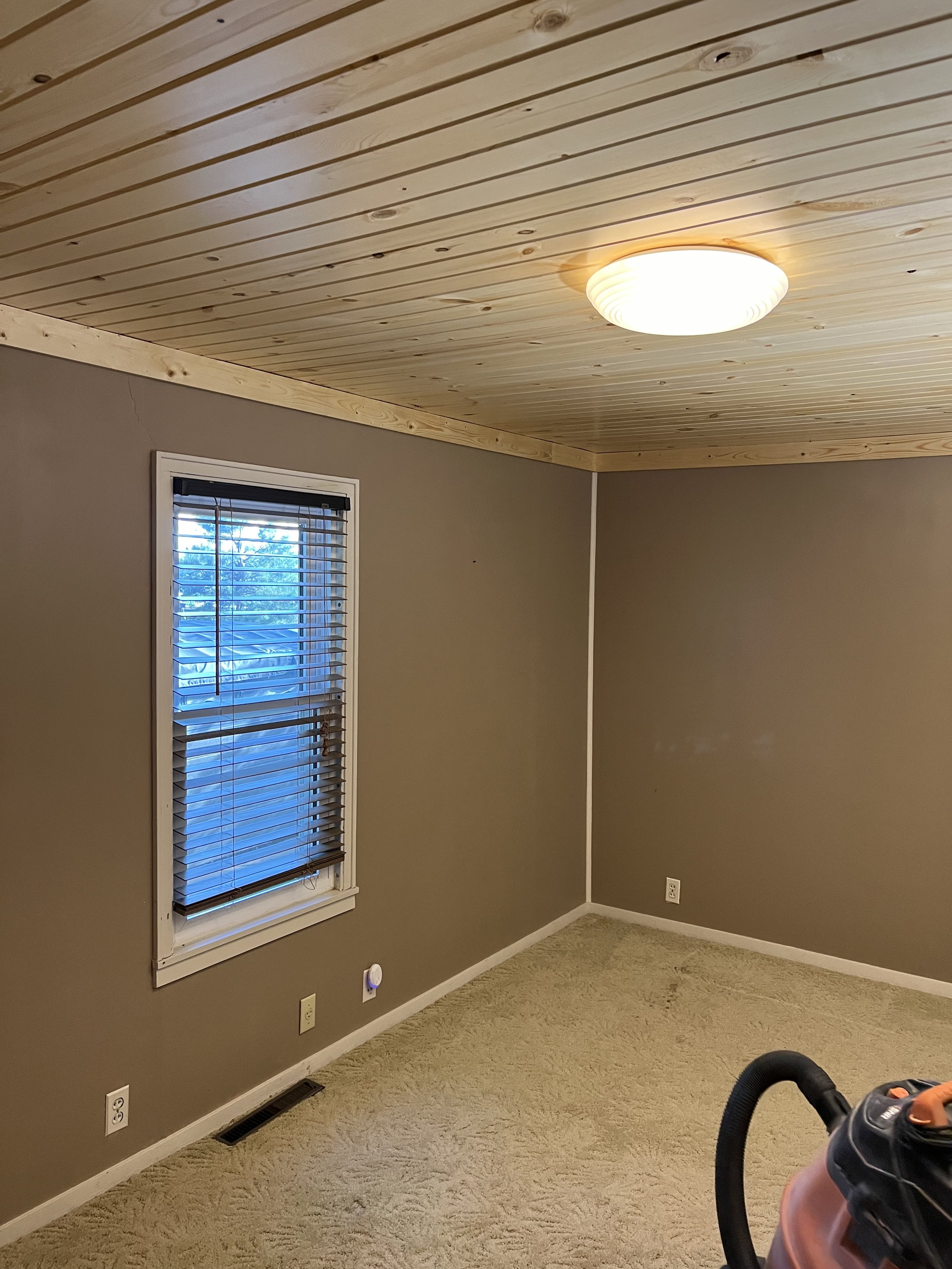 empty room with wooden ceiling and beige carpet, a single window with blinds, a ceiling light, and a vacuum cleaner in the foreground