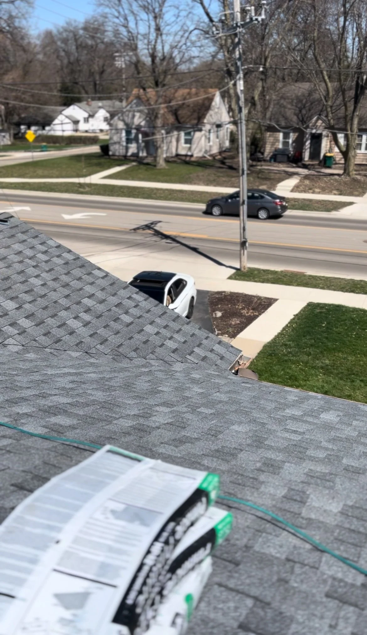 View of a street with houses, cars, and power lines from a rooftop with gray shingles and roofing materials in the foreground.