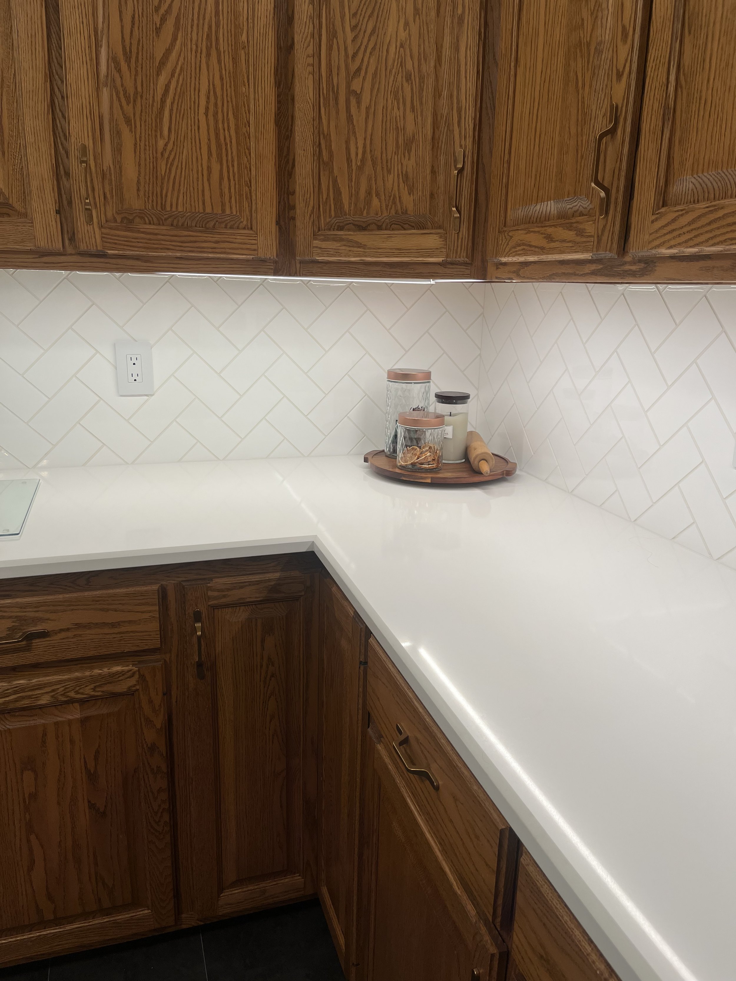 Modern kitchen counter with wooden cabinets, white quartz countertop, and herringbone tile backsplash. A tray with jars and a rolling pin is placed on the counter.