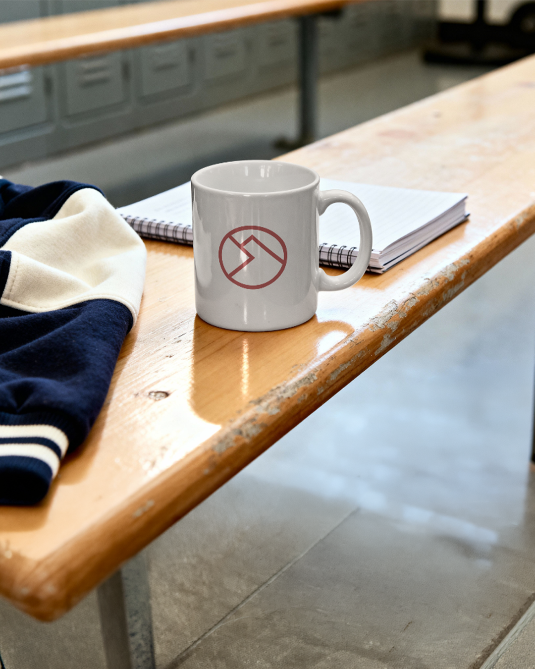 White coffee mug featuring the Bellavita logo sitting on a locker room bench near a sweater and notebook.