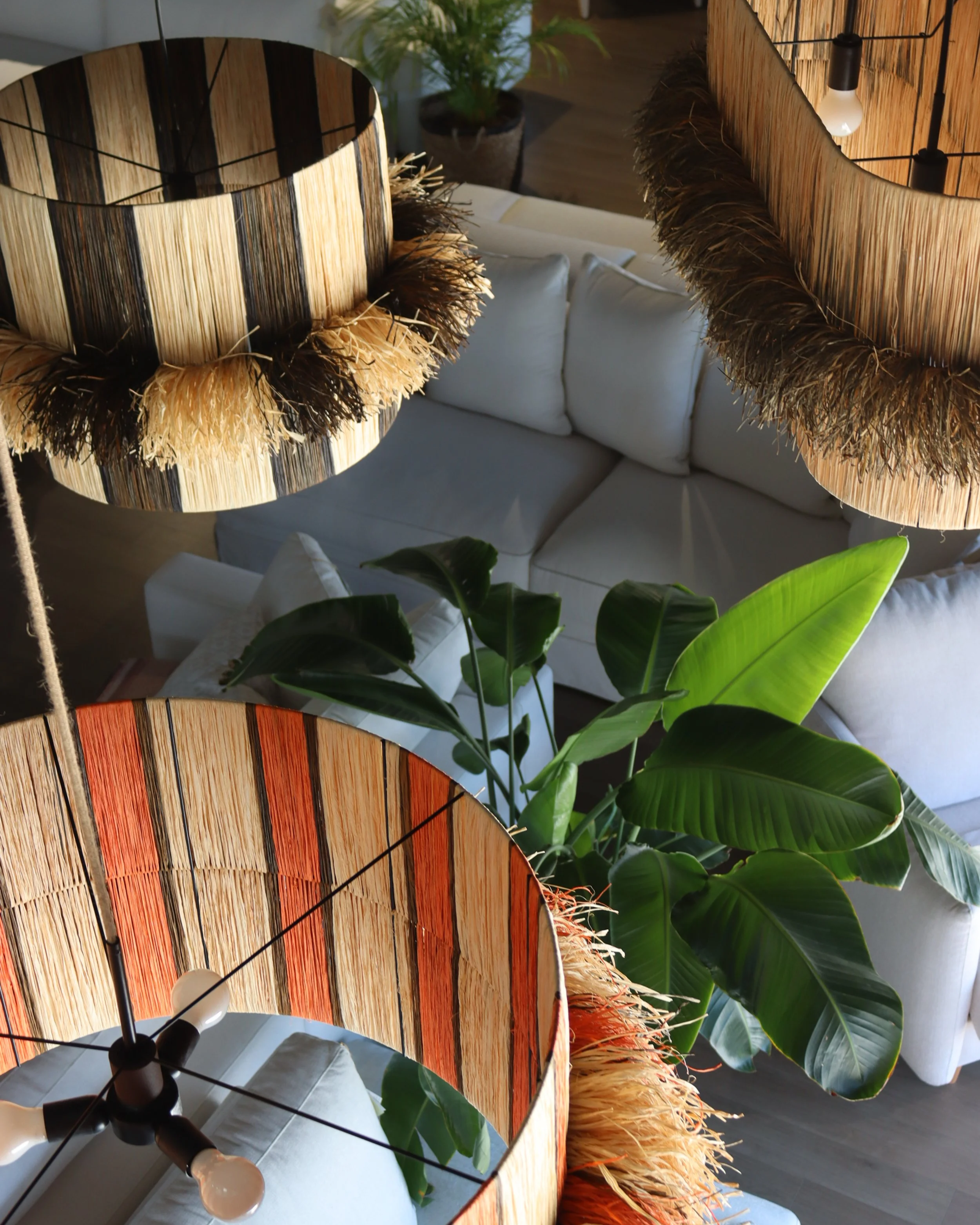 Lámparas de techo de madera con borde de fibras naturales, luz encendida, vista desde arriba en una sala con sofá blanco, planta grande con hojas verdes.