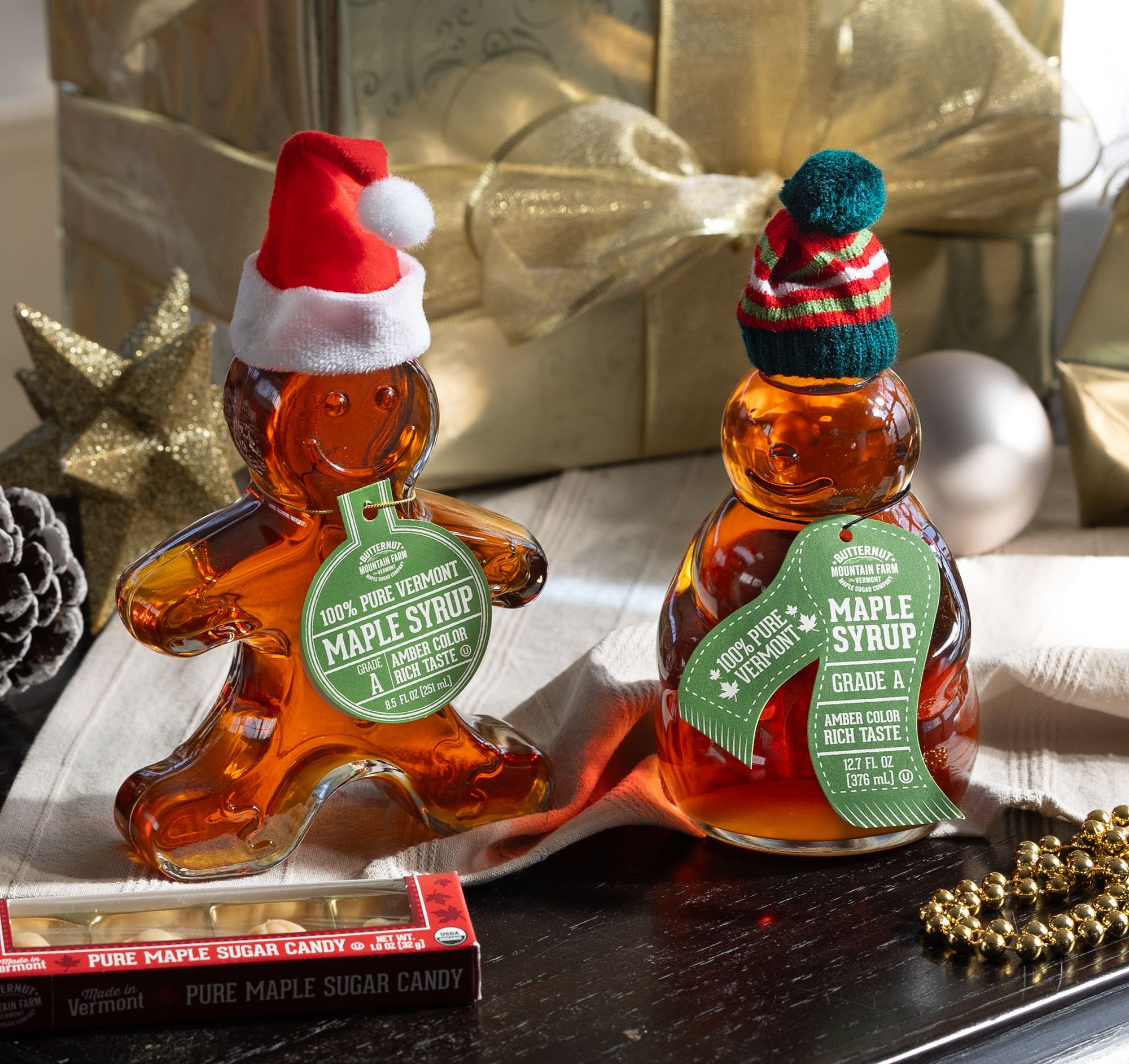 A gingerbread man shaped bottle of maple syrup and a snowman shaped bottle of maple syrup posed in front of a gold present with a gold bow.