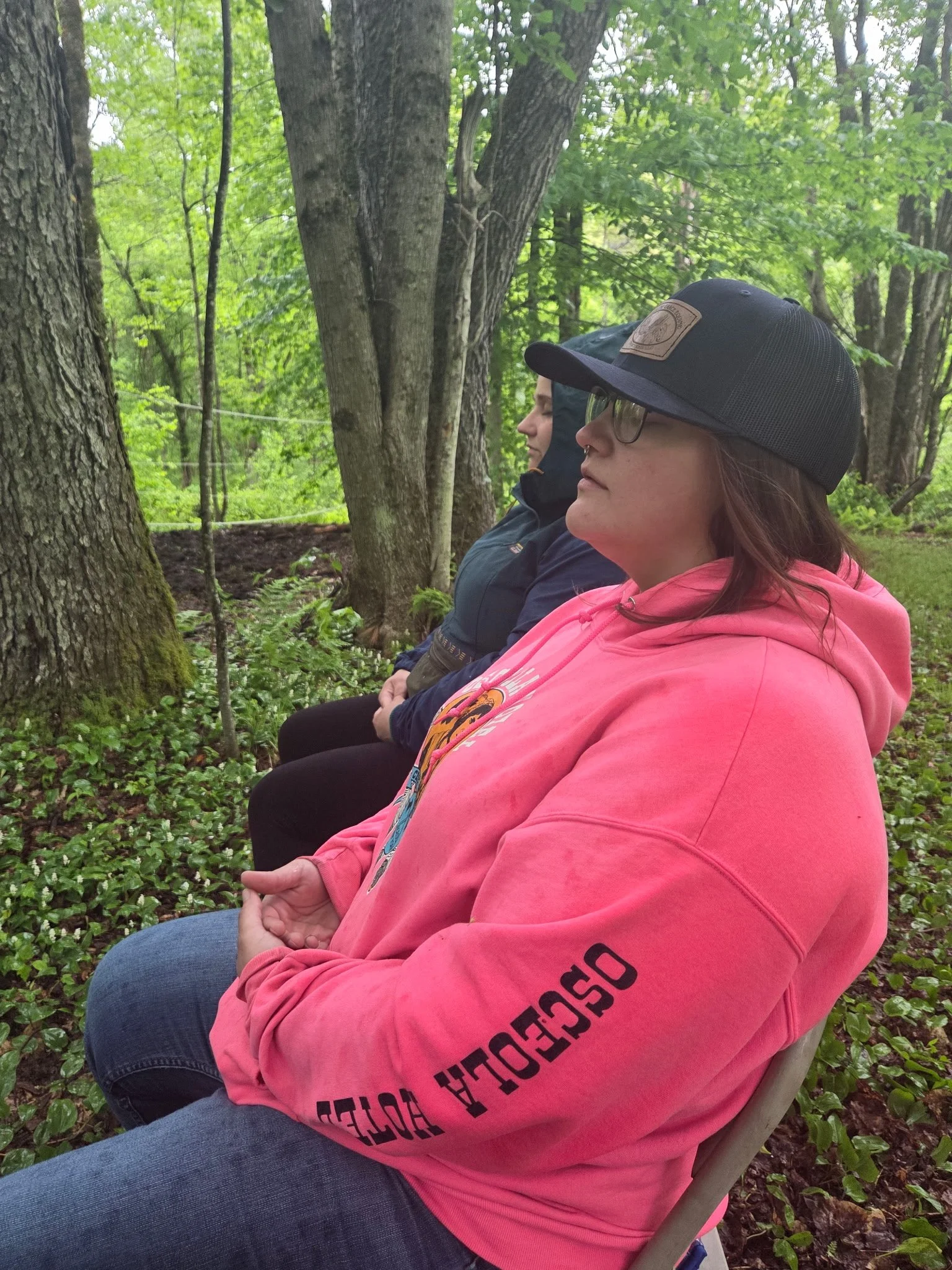 Two women sitting outdoors in a forest, seated in a row with their eyes closed, hands resting on their laps, meditating or relaxing.