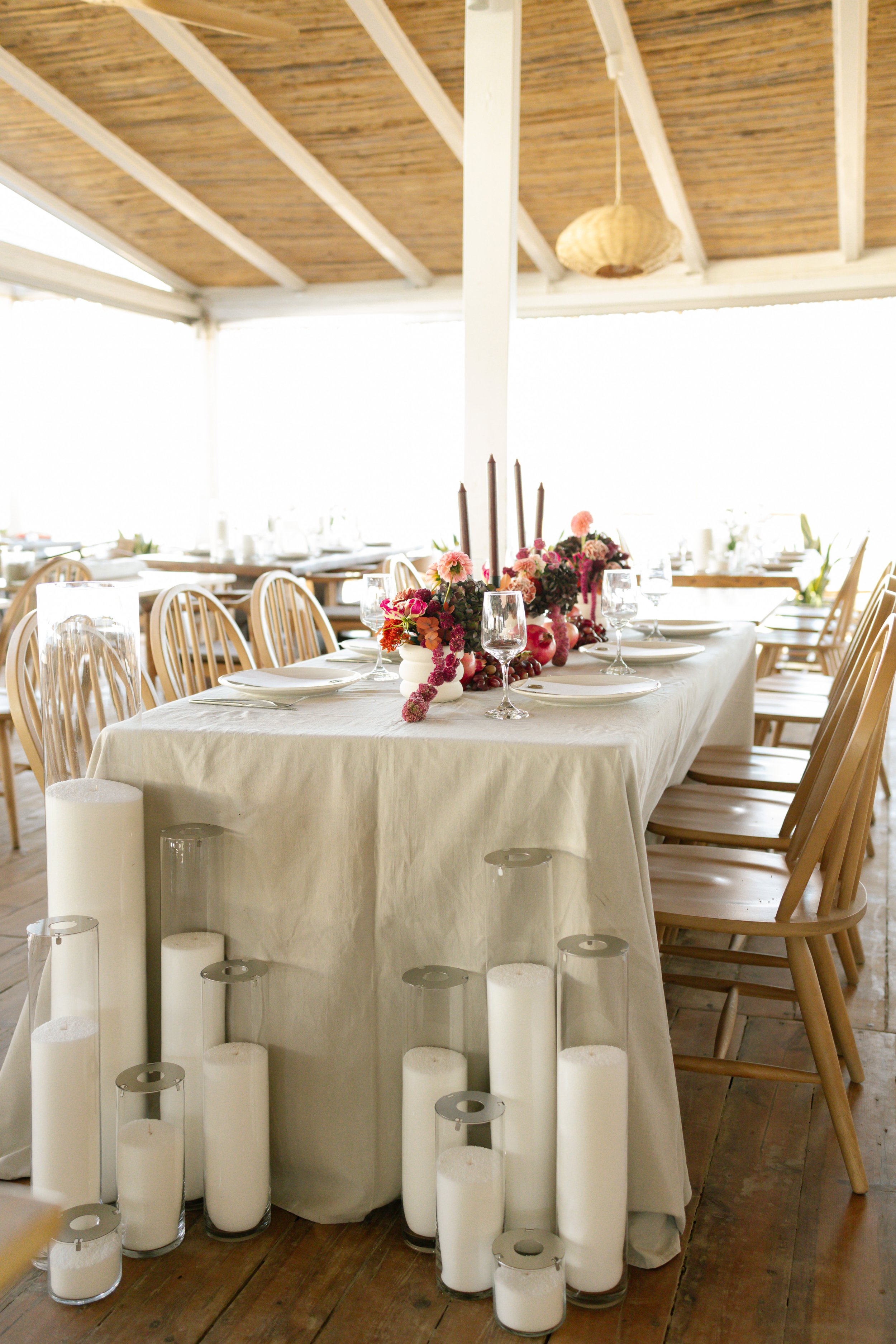 Elegant seaside wedding table setup in Athens, featuring a white tablecloth, lush floral centerpiece, candles, and rustic wooden chairs. The romantic coastal setting, perfect for a luxury destination wedding, creates a timeless and sophisticated atmo