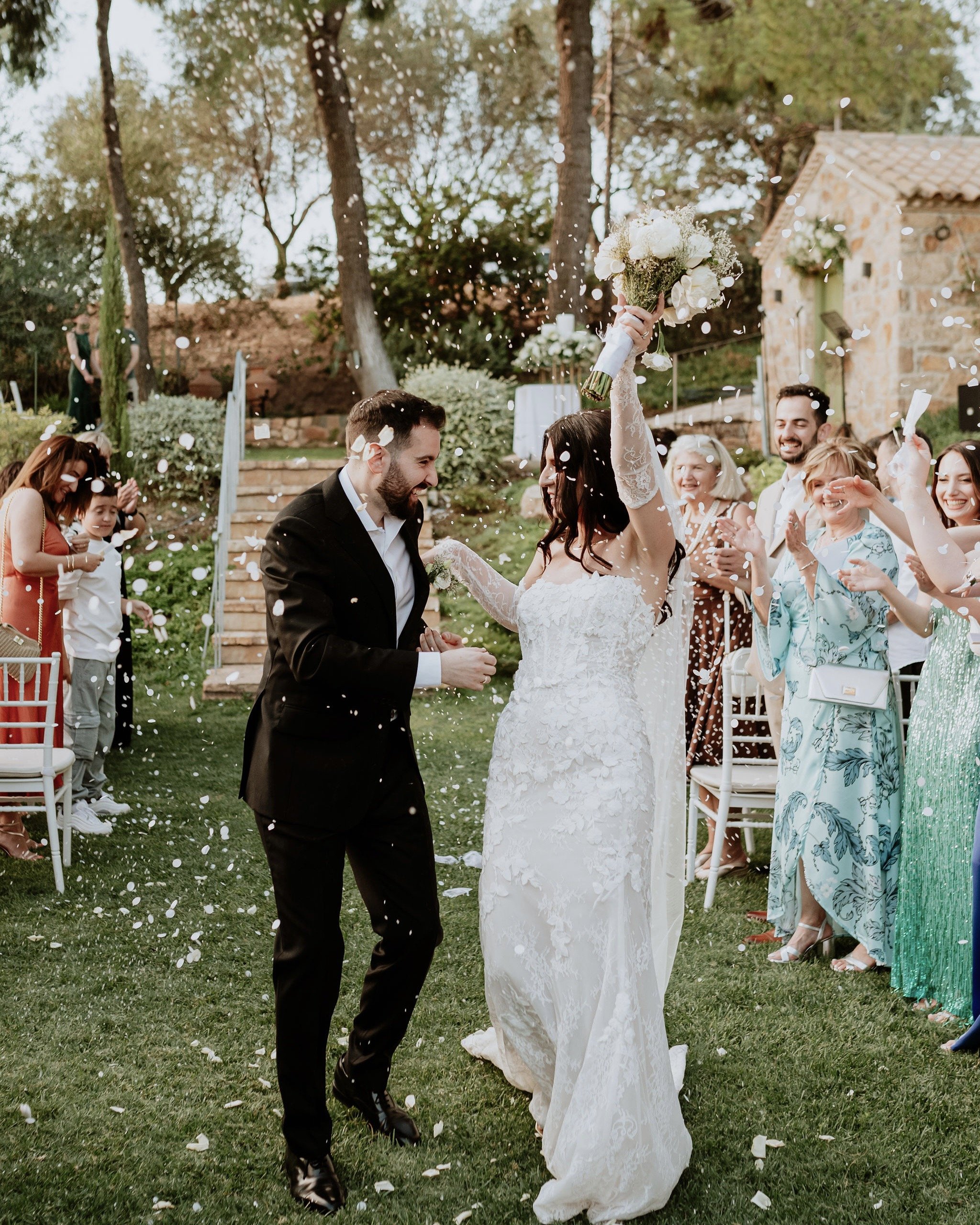 Bride and groom celebrating their wedding exit outdoors in Greece, surrounded by guests tossing confetti. Lush greenery and natural surroundings create a joyful and intimate atmosphere, perfect for a timeless, romantic destination wedding in Greece.