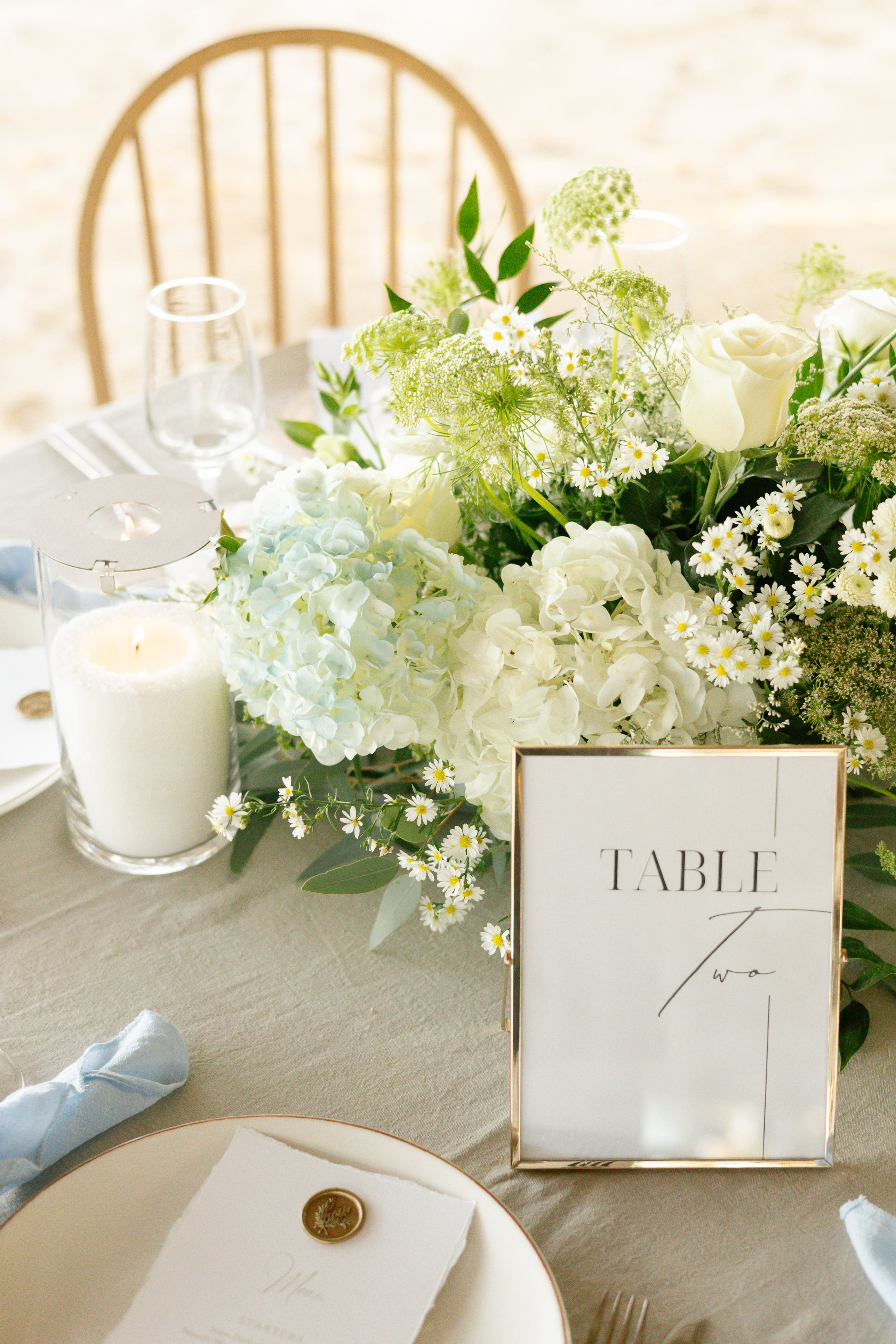 Elegant table setting with white and green floral centerpiece, table number card, candle, glassware, and a white plate with menu on a light tablecloth.