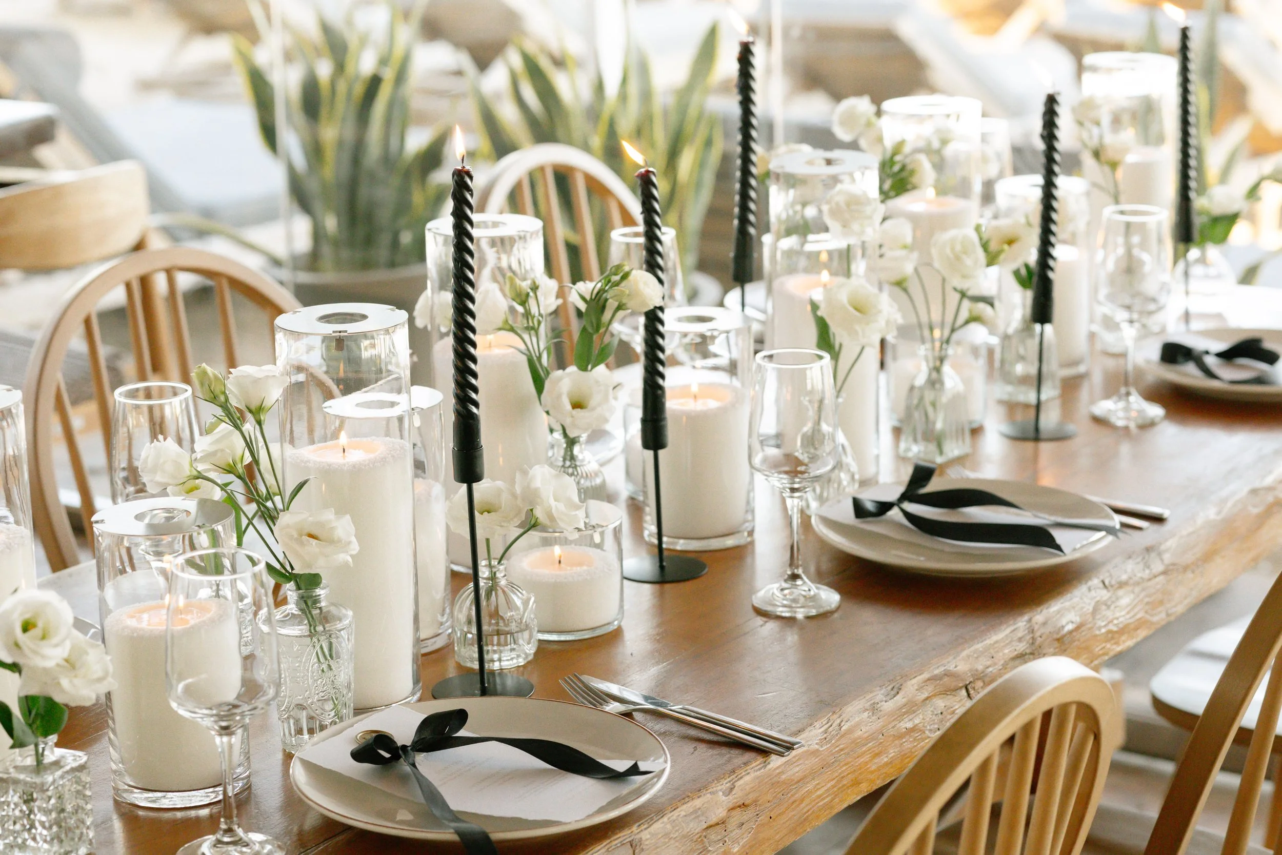 Elegant wedding table setting in Greece featuring white candles, black twisted tapers, white flowers in vases, glassware, and plates with black ribbons. Wooden chairs and table create a sophisticated and timeless atmosphere for a luxury wedding