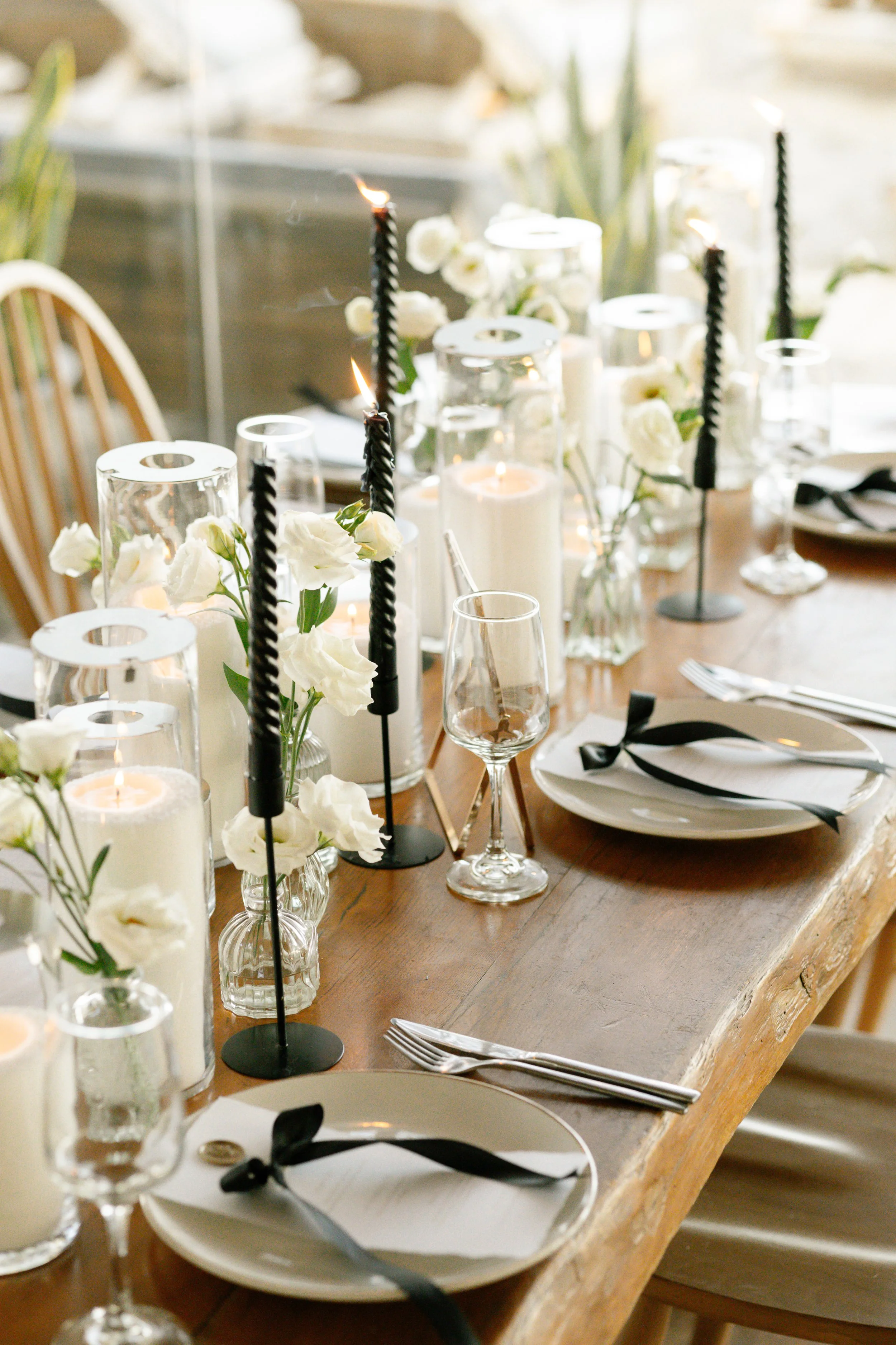 Elegant dining table setting for a luxury wedding in Greece, featuring black candles, white flowers, glass candle holders, plates with black ribbons, and wine glasses on a wooden table. Perfect for sophisticated, timeless wedding receptions in Athens