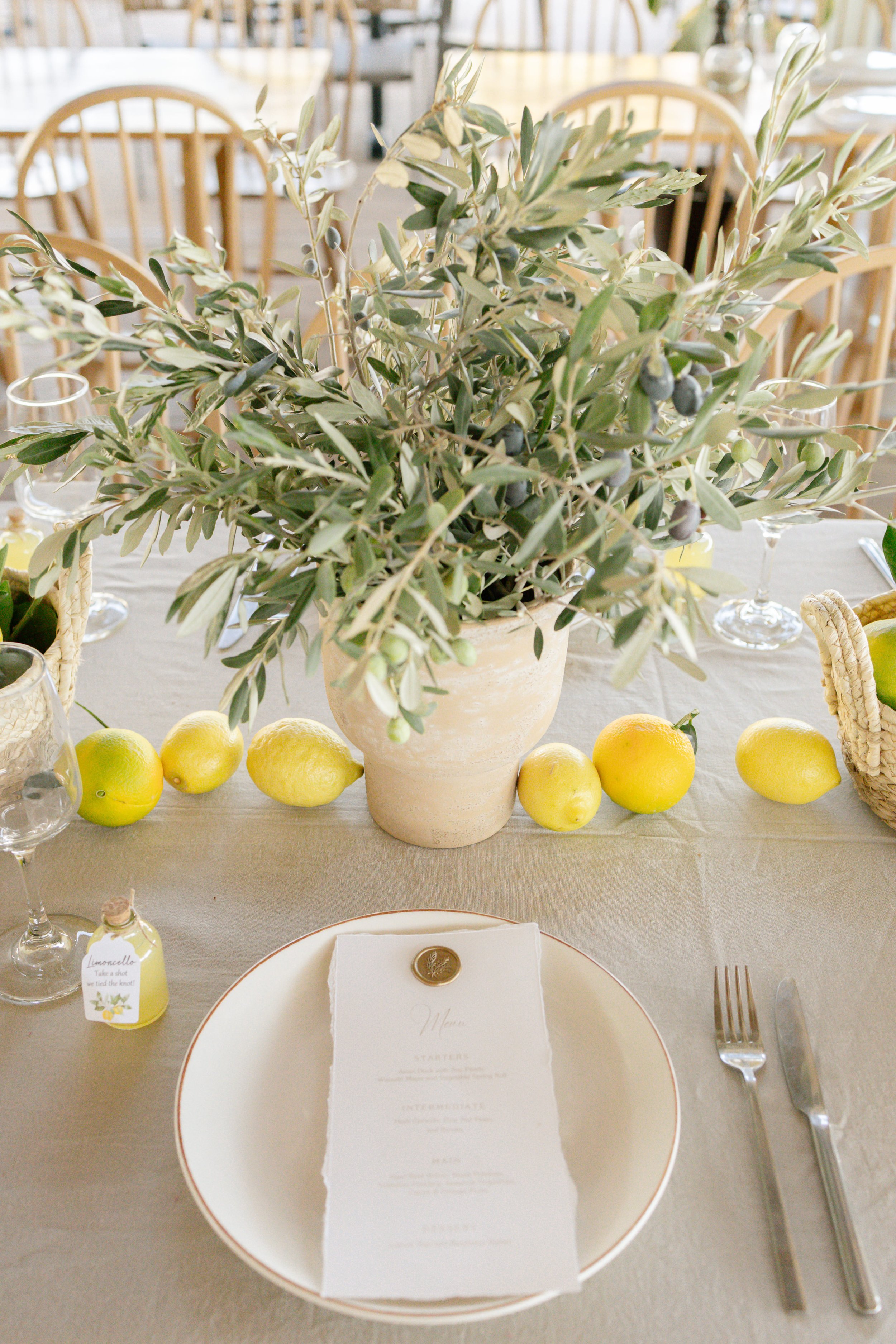 Charming wedding table setup in Greece with a white plate, elegant menu card, and cutlery. Olive branches in a vase create a rustic centerpiece, while lemons and a bottle of 'Limoncello' bring a refreshing Mediterranean flair.