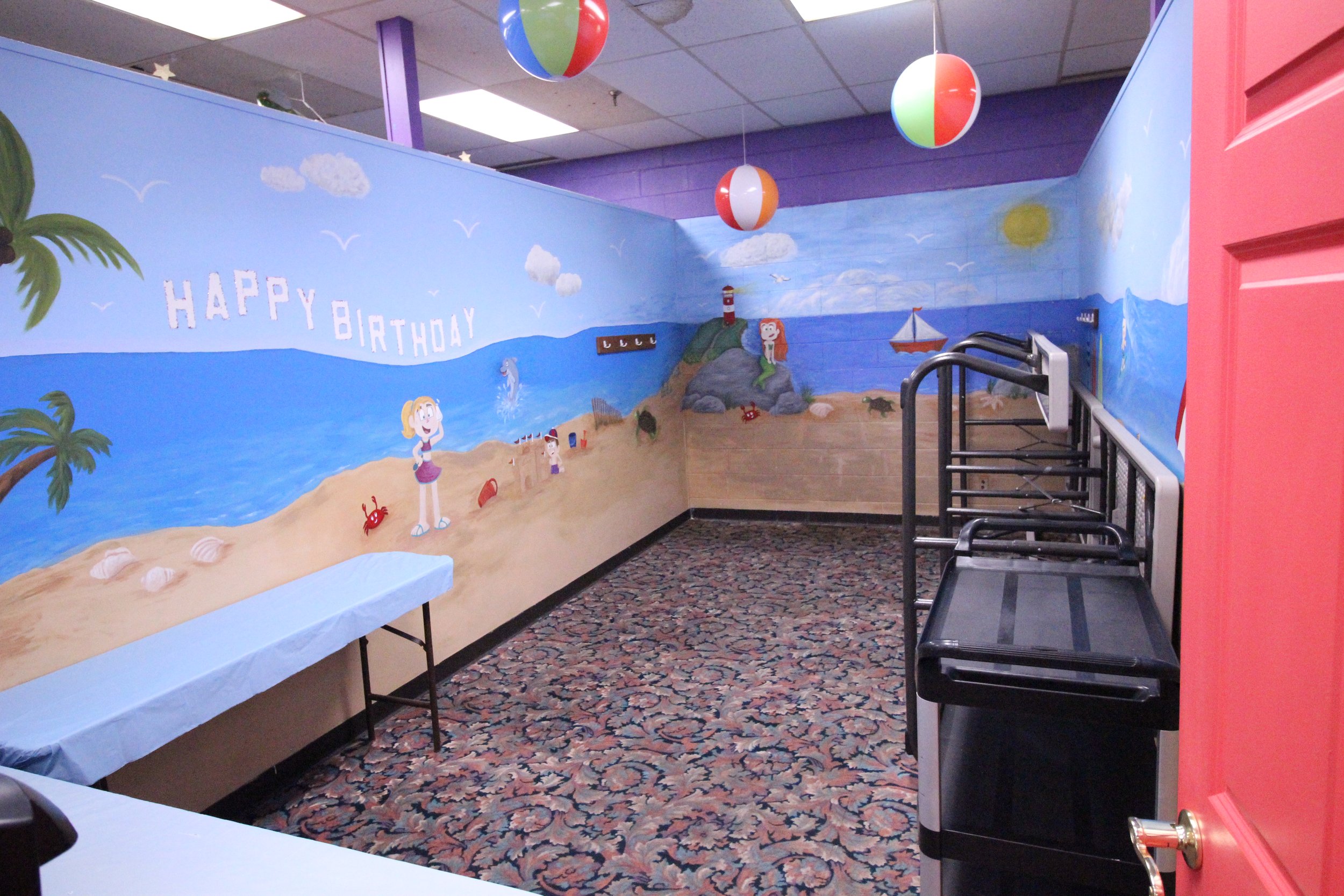 Room with beach-themed mural and "Happy Birthday" message, decorated with beach balls and tables.