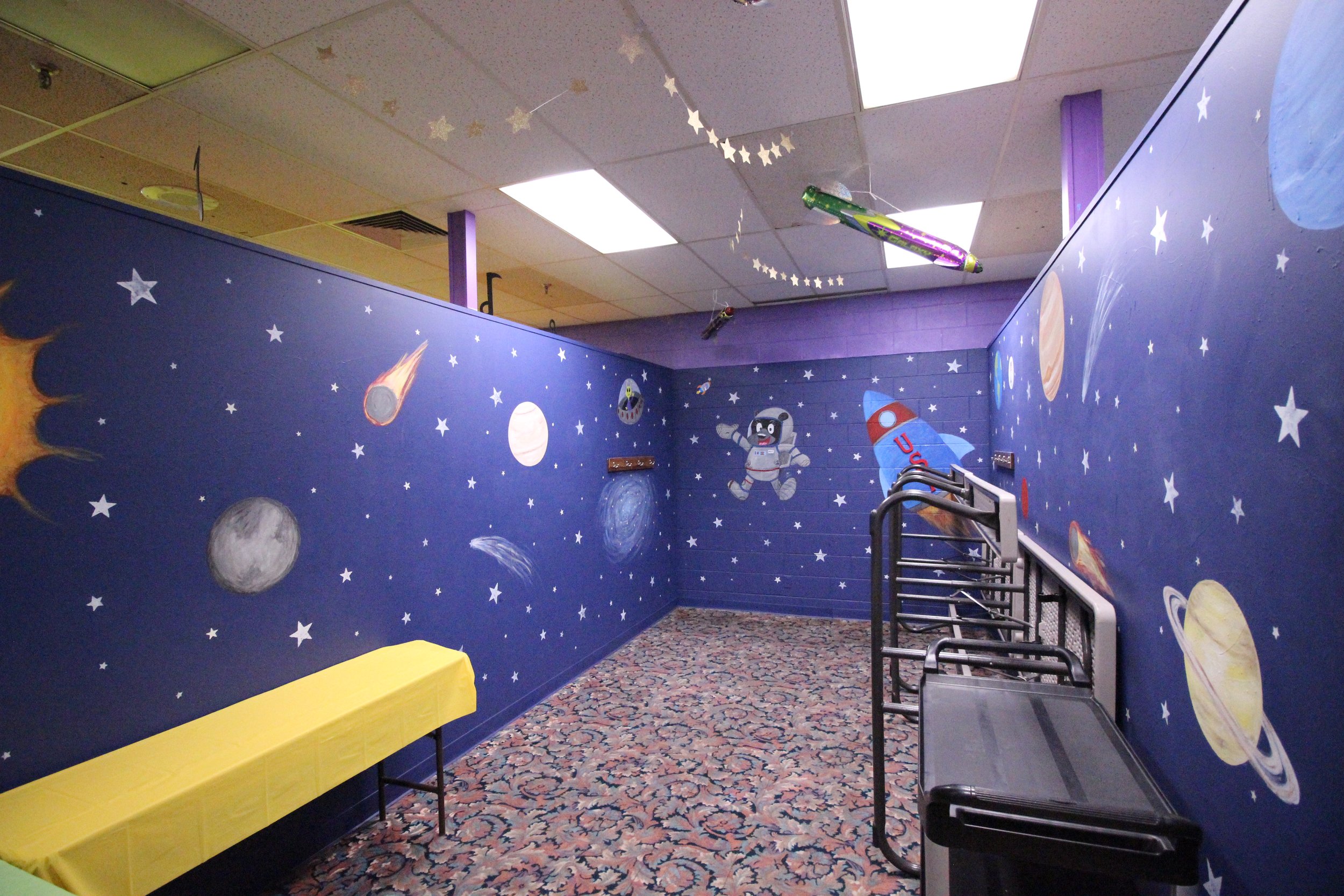 Room with space-themed wall mural, featuring planets, stars, and a cartoon astronaut. Yellow tablecloth-covered tables and stacked chairs. Ceiling decorations include stars and colorful hanging items.