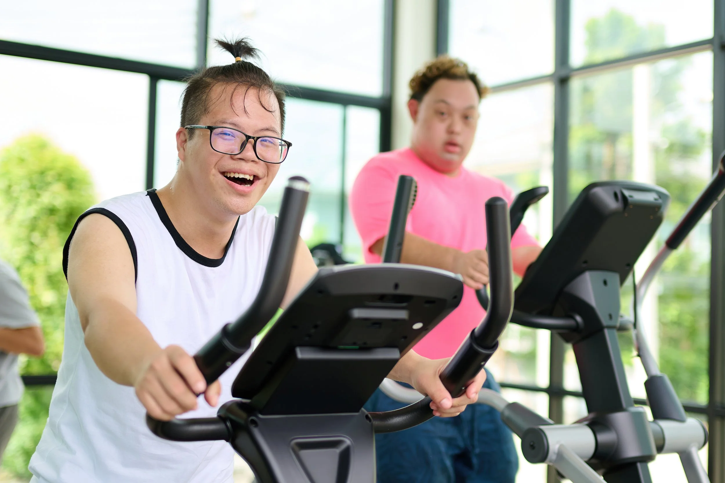 Two young men exercising on elliptical machines in a bright indoor gym with large windows and green outdoor scenery.