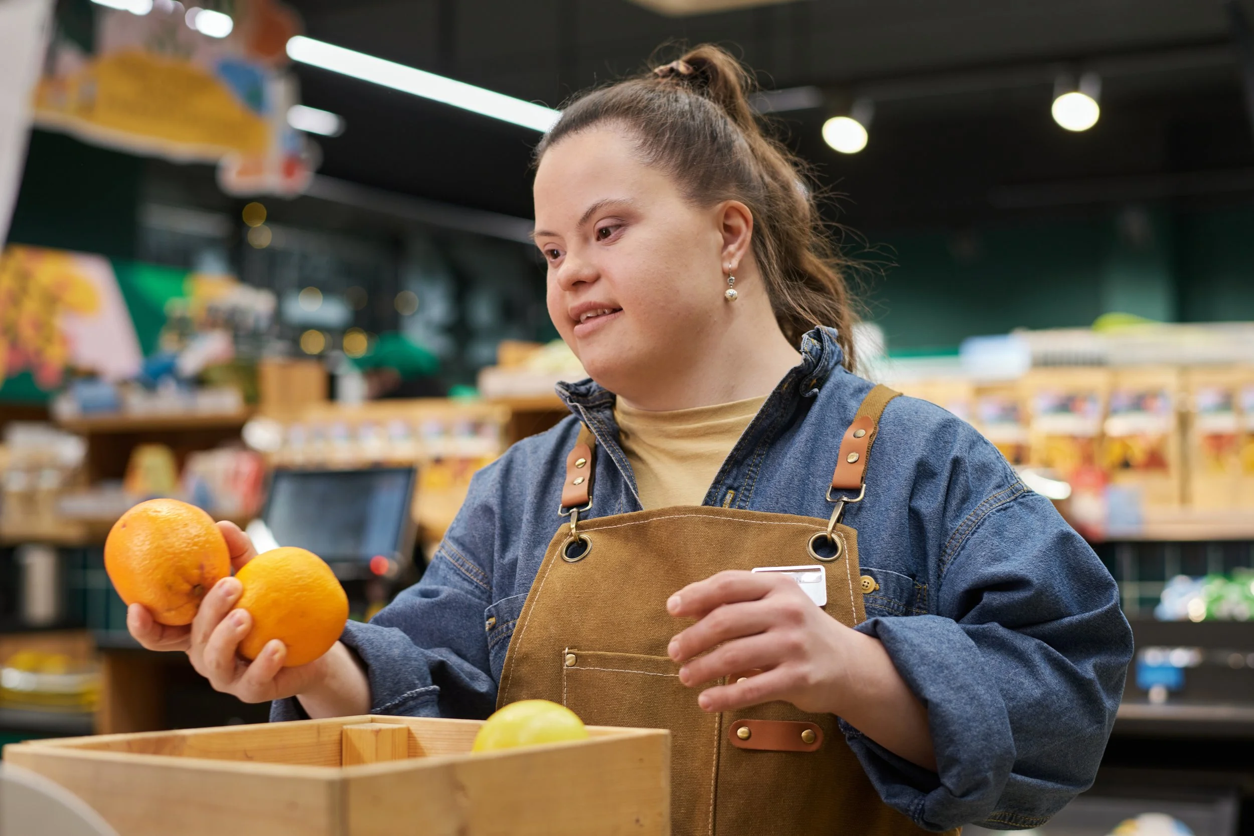A woman working at a grocery store checkout counter, holding oranges in one hand and a credit card in the other, wearing a denim jacket and a tan apron.