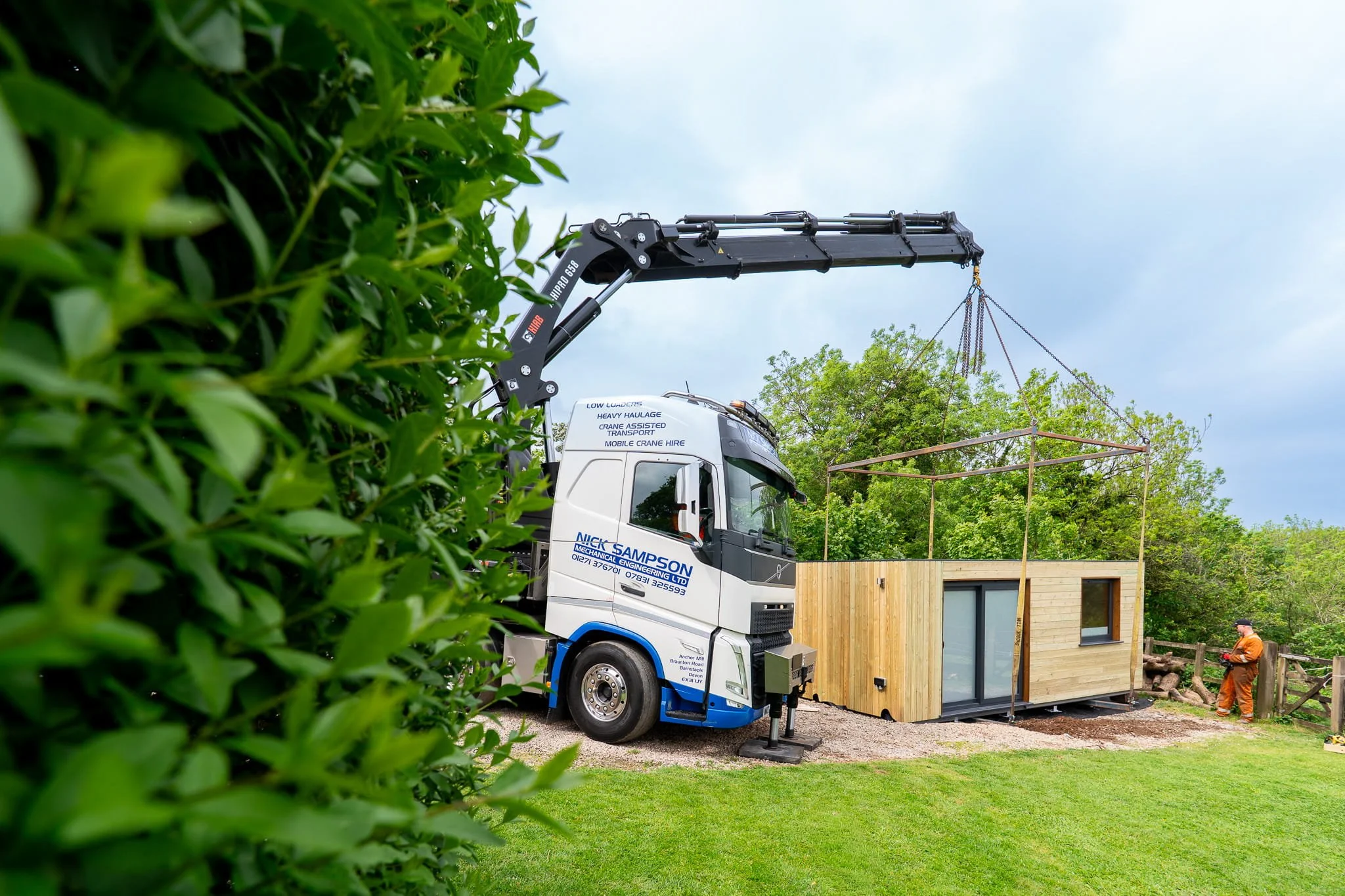 A construction site with a crane truck lifting a garden room, with workers and green bushes in the foreground and trees in the background.