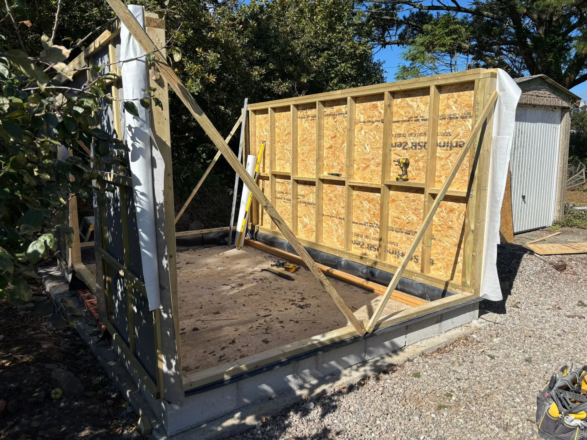 Construction of a small wooden shed on a concrete foundation on a gravel surface, with partially built walls and construction tools nearby.