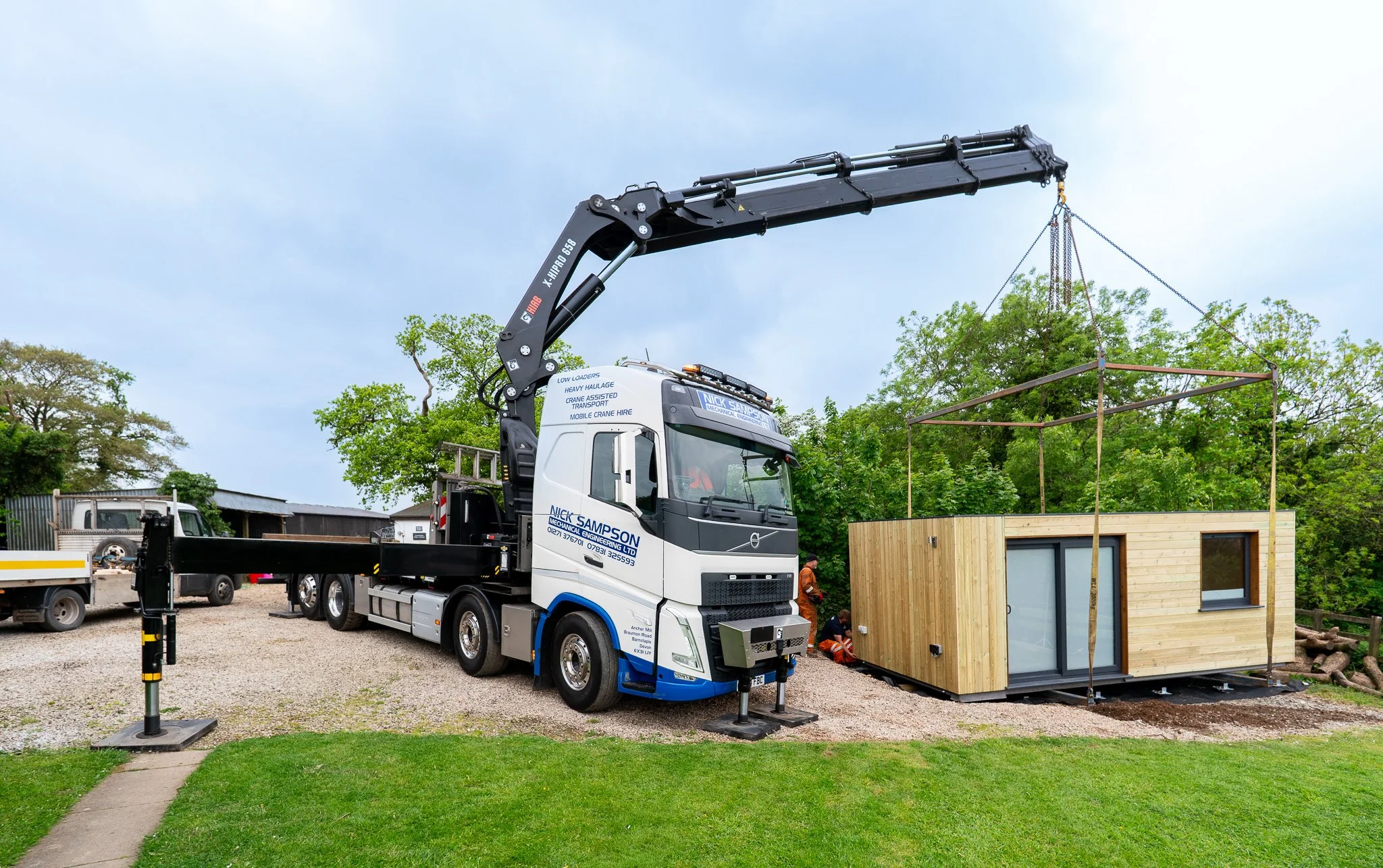 A large white truck with a crane lifting a cabin. The scene is outdoors with green trees and a cloudy sky in the background.