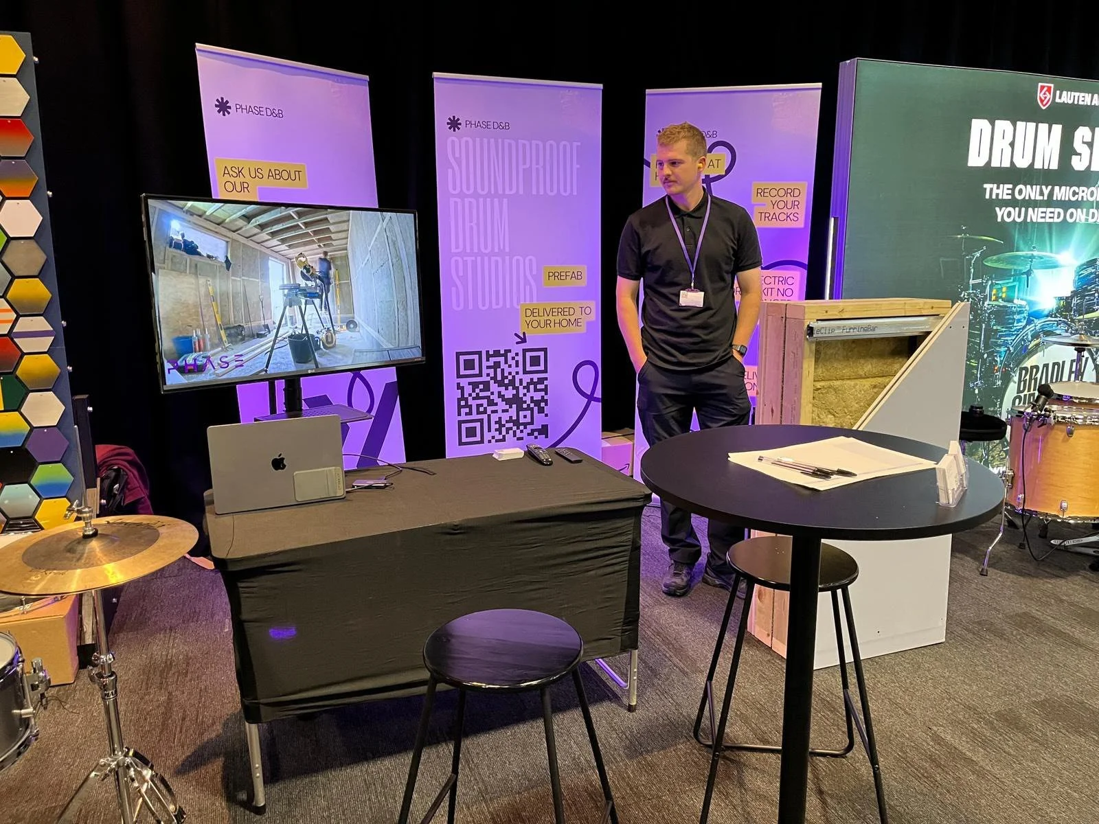 At a trade show booth, a man in a black shirt and pants stands with hands in pockets near a display with banners and a large monitor showing construction inside a wooden room. The booth has promotional materials, banners with QR codes, drum parts, an