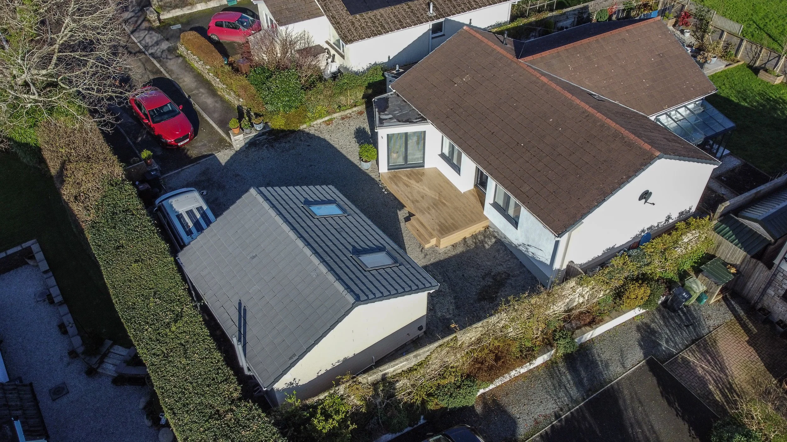 Aerial view of a residential backyard showing a house with a wooden deck, a smaller building with skylights, parked cars, and surrounding trees and fences.