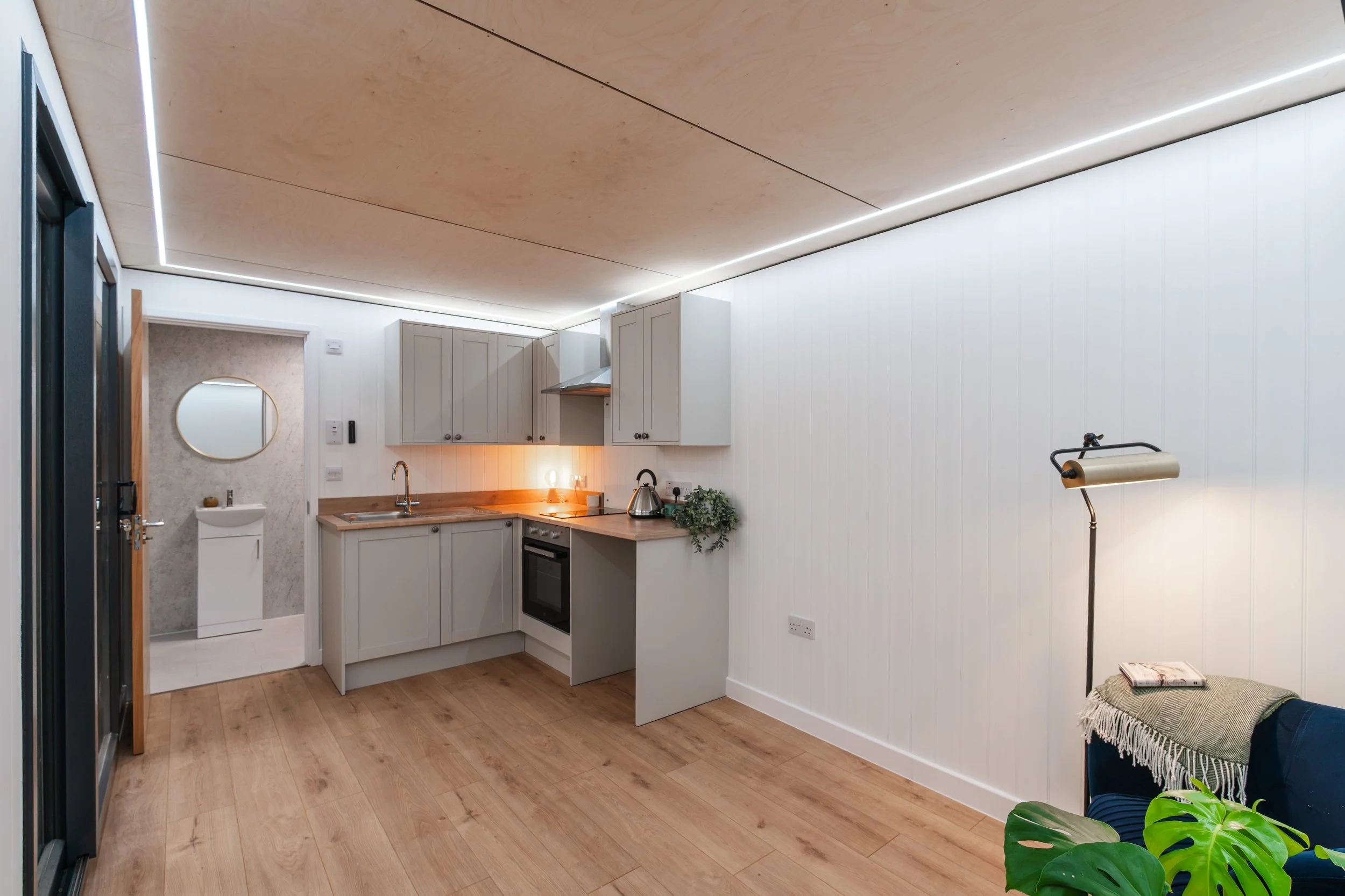 A modern kitchen with white cabinets, a wooden countertop, and a stove. There is a small circular mirror on the wall near a door leading to a bathroom. The ceiling has wooden panels with built-in LED lighting. A floor lamp and some plants are visible