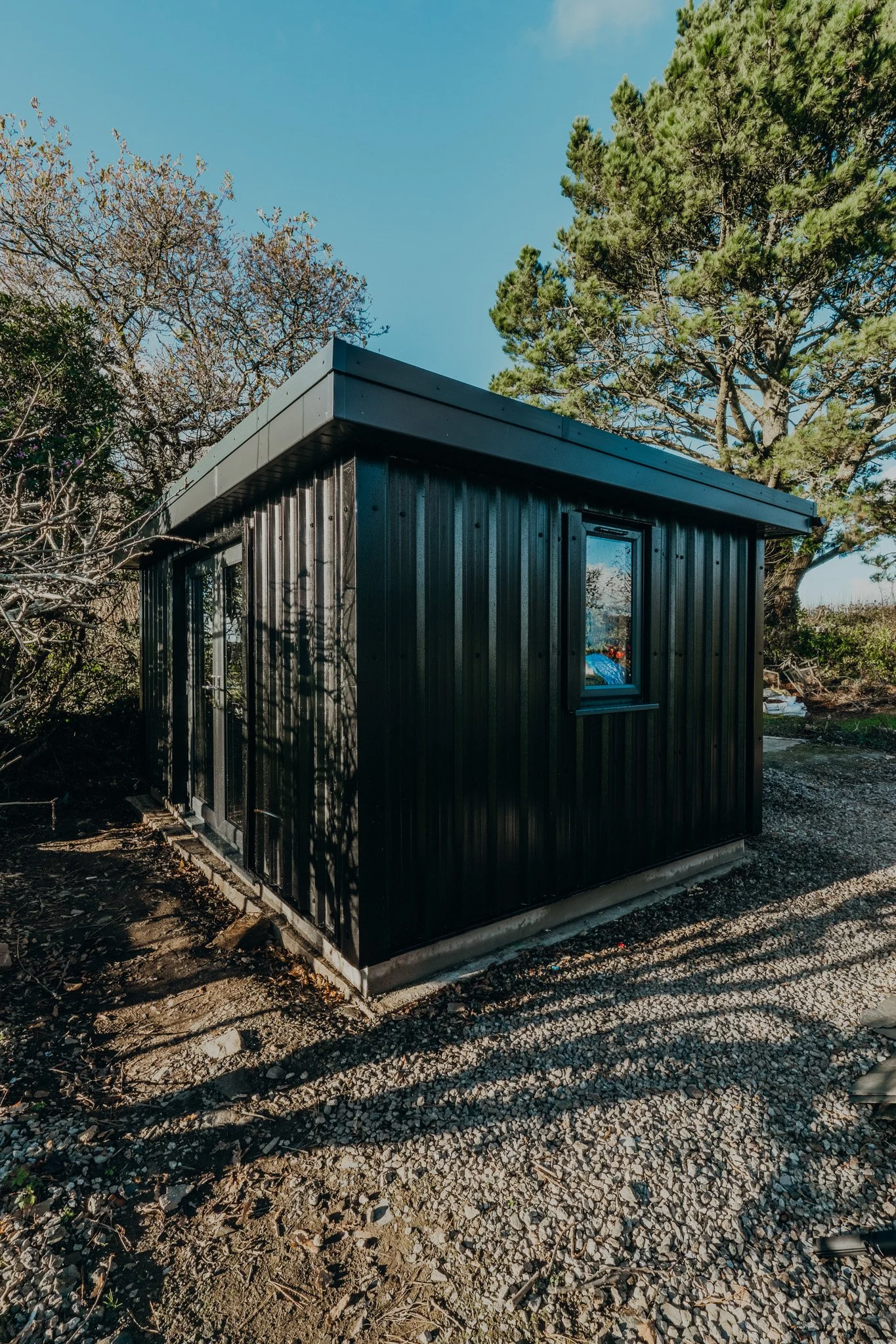 A small black metal shed with a flat roof, situated on a gravel surface, surrounded by trees and bushes under a clear blue sky.