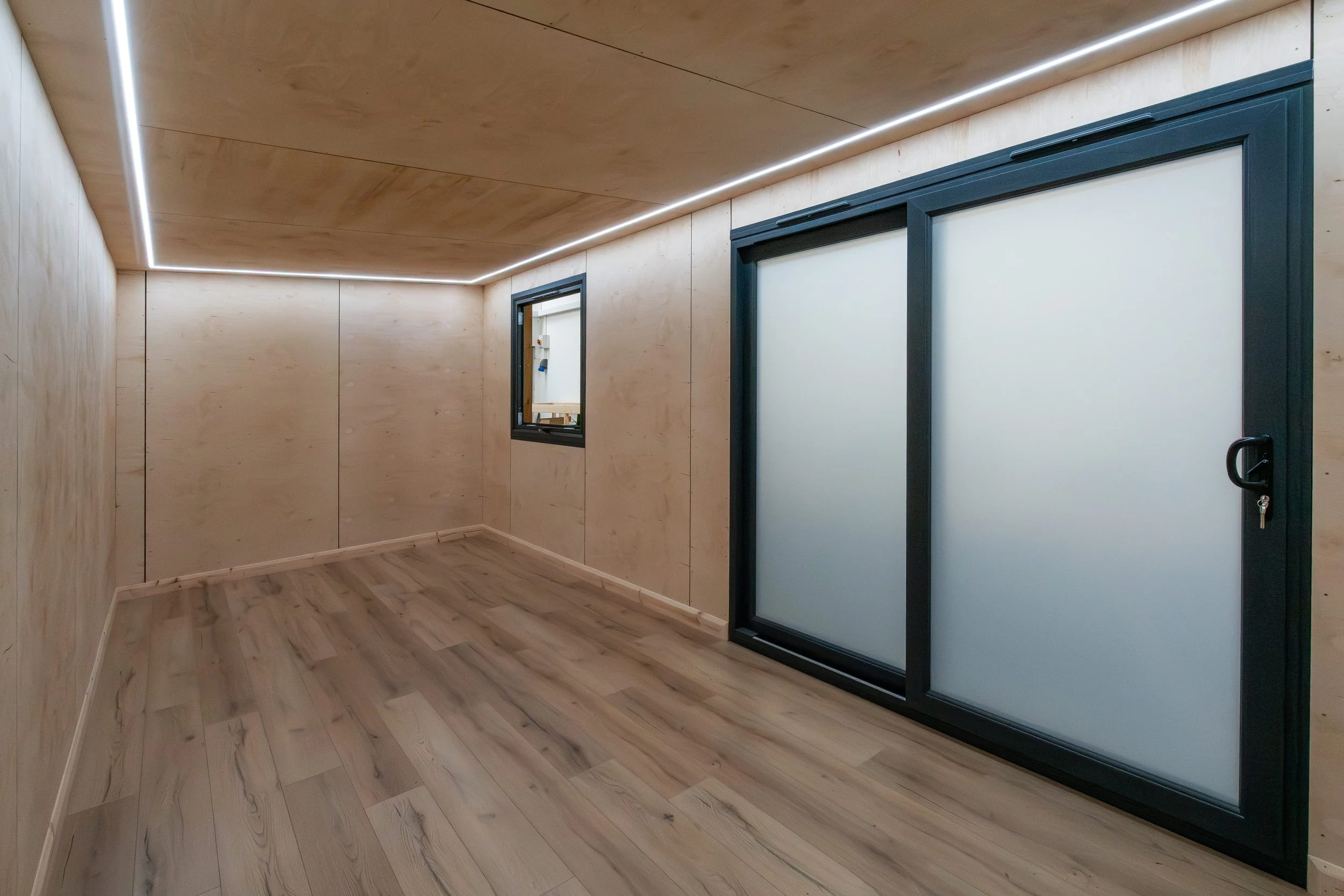 An empty garden room with wooden walls and flooring, a window on one wall, and sliding doors with frosted glass panels and black frames.