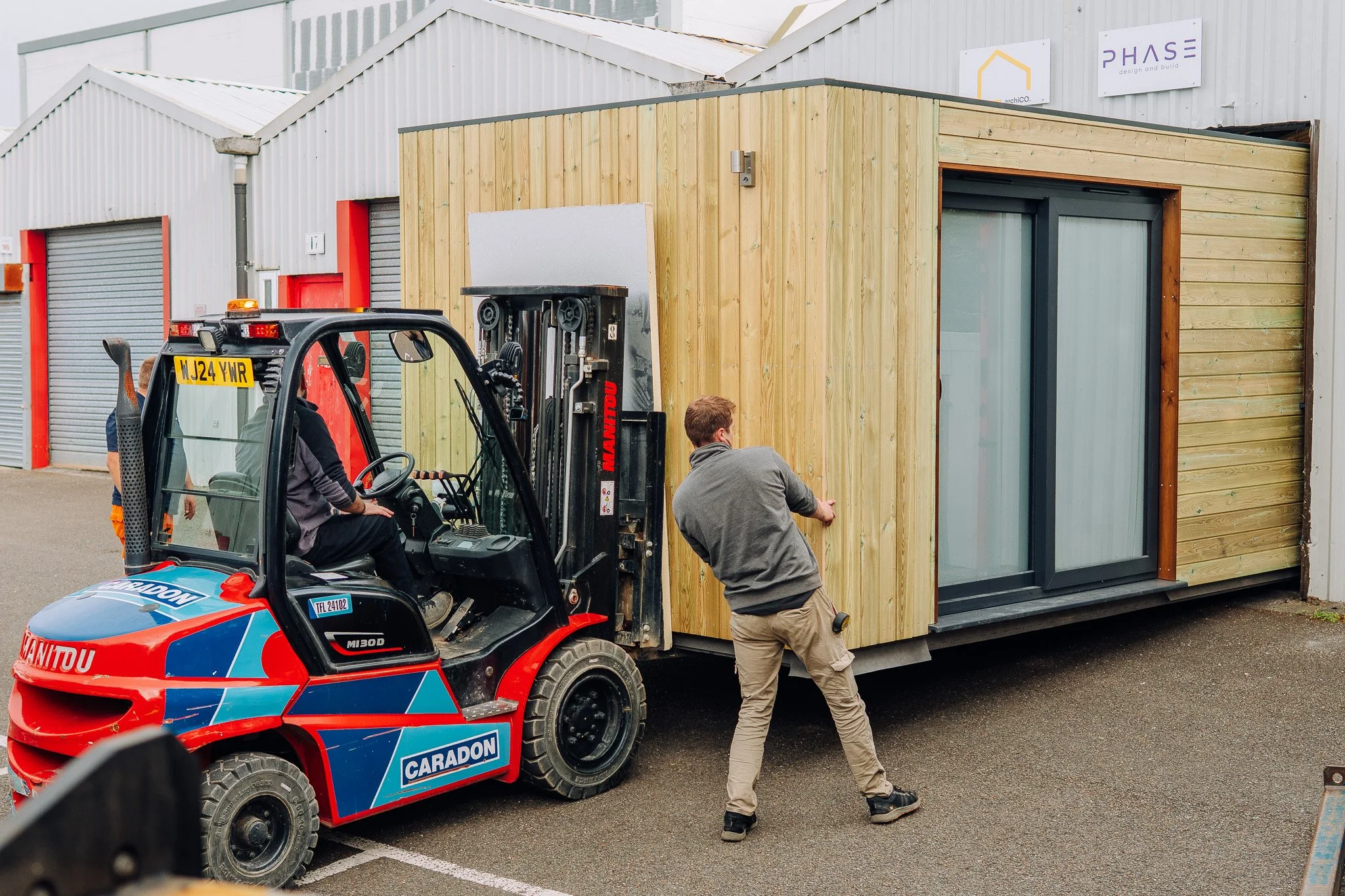 Two men are working with a forklift to position a garden room cabin outside a warehouse. The warehouse has a metal exterior with signs reading 'archiCO' and 'PHASE design and build'. The scene is set in a parking lot.