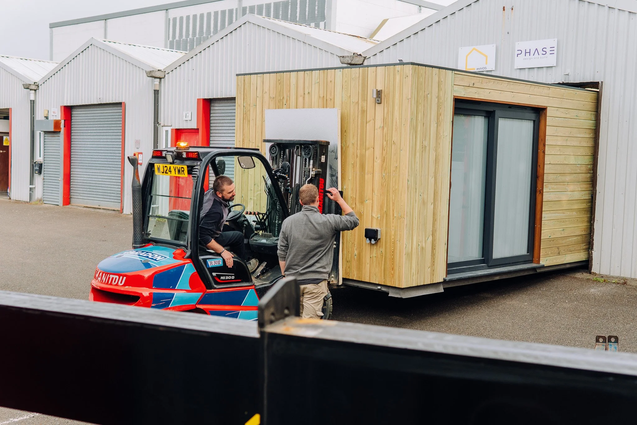 Two men are working with a forklift truck, moving a garden room cabin or container, with multiple retail units or warehouses in the background.