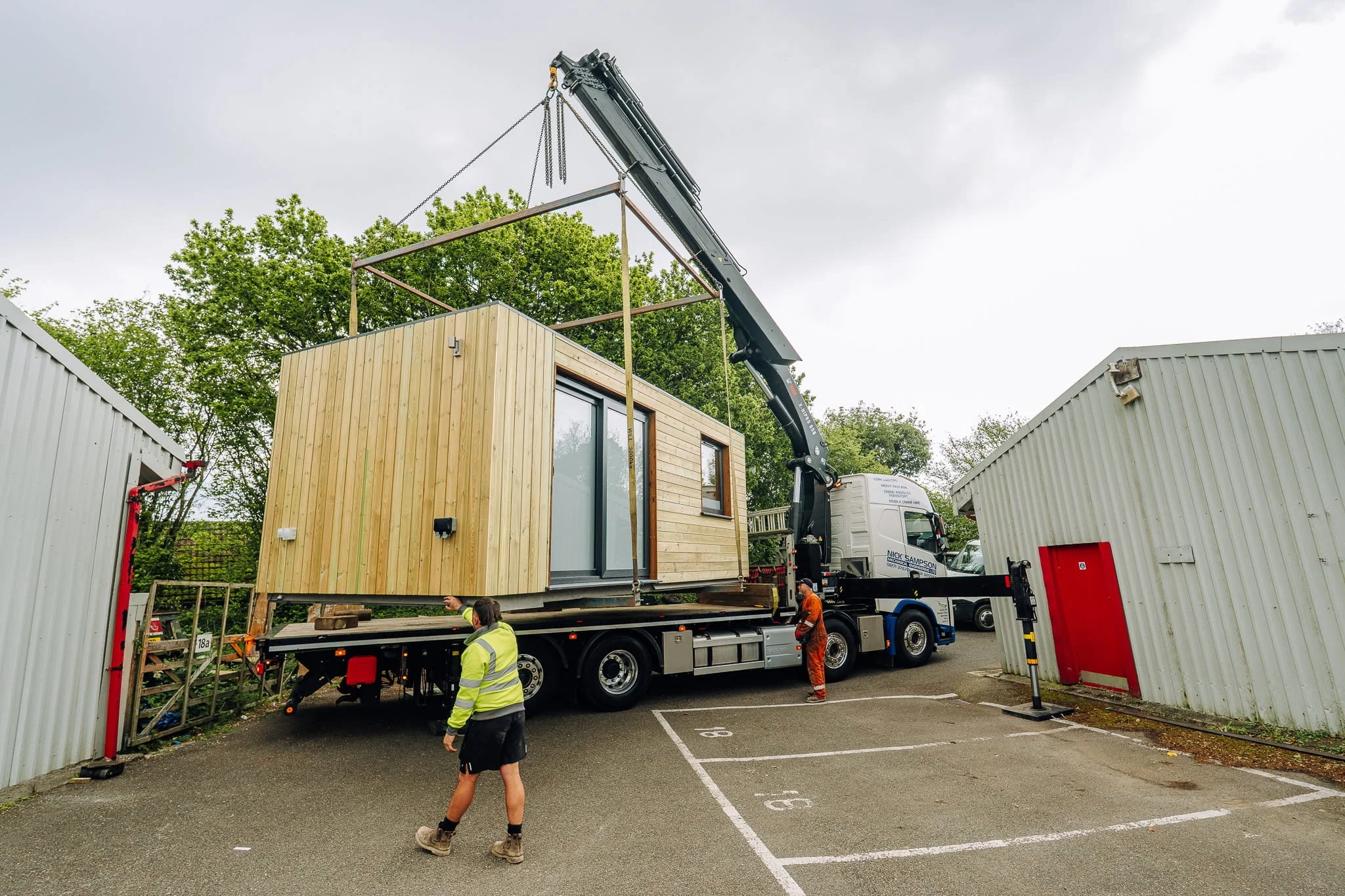A large crane lifting a small phase garden room house onto a flatbed truck at a construction site, with workers overseeing the process.