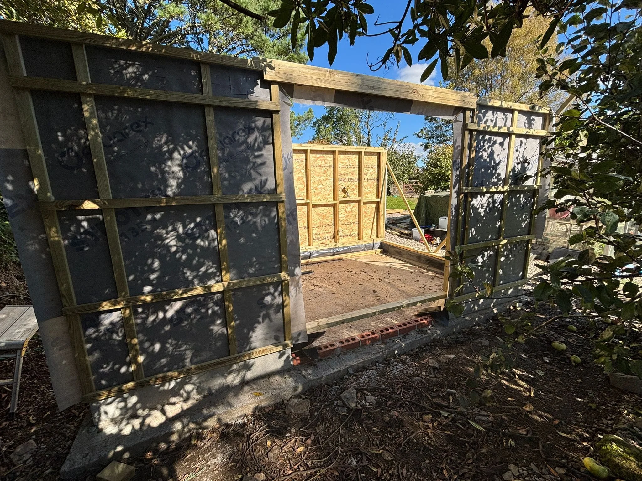 Construction of a small wooden structure or shed outdoors with framing and partial wall panels in progress, surrounded by trees and plants.