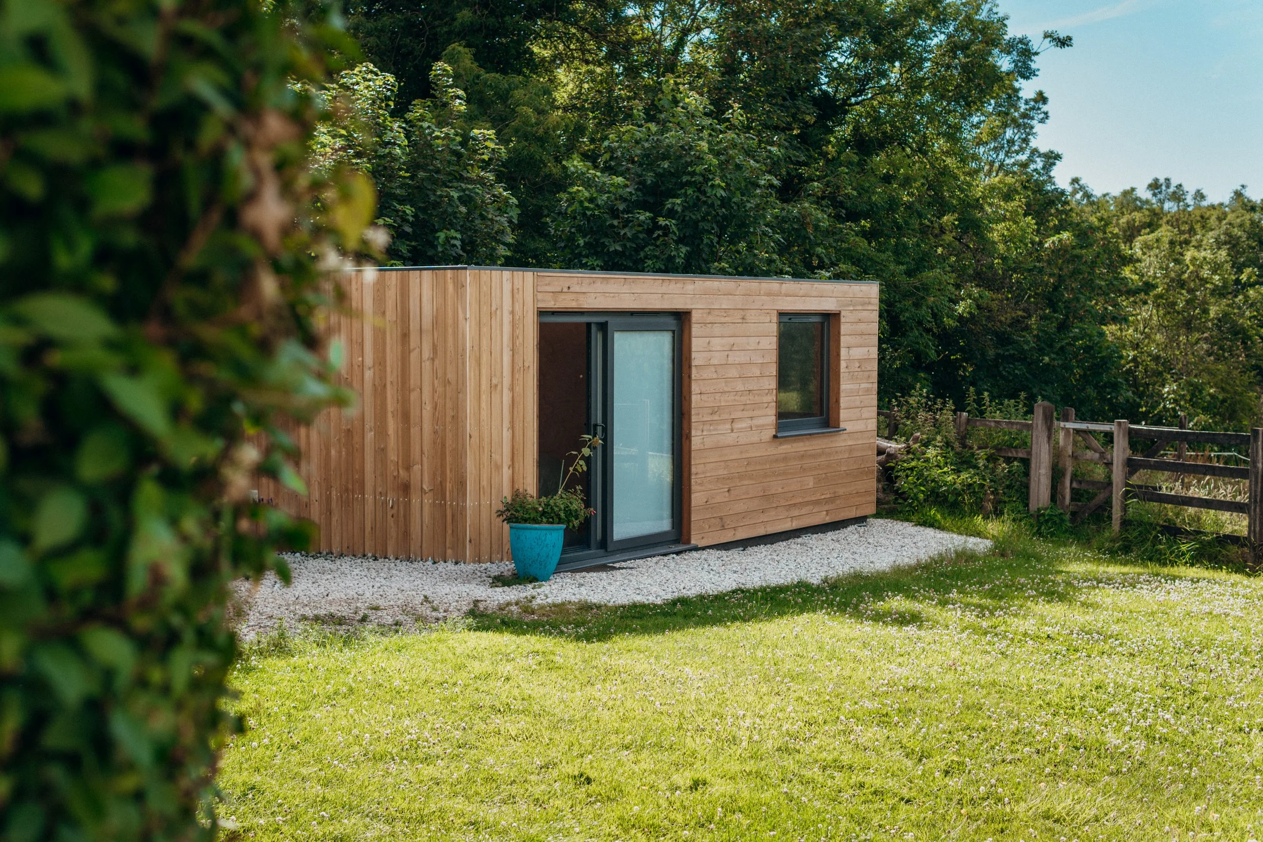 A wooden tiny house with a glass door and a small window, situated on a gravel area, surrounded by green grass and trees, with a blue pot containing a small plant in front.