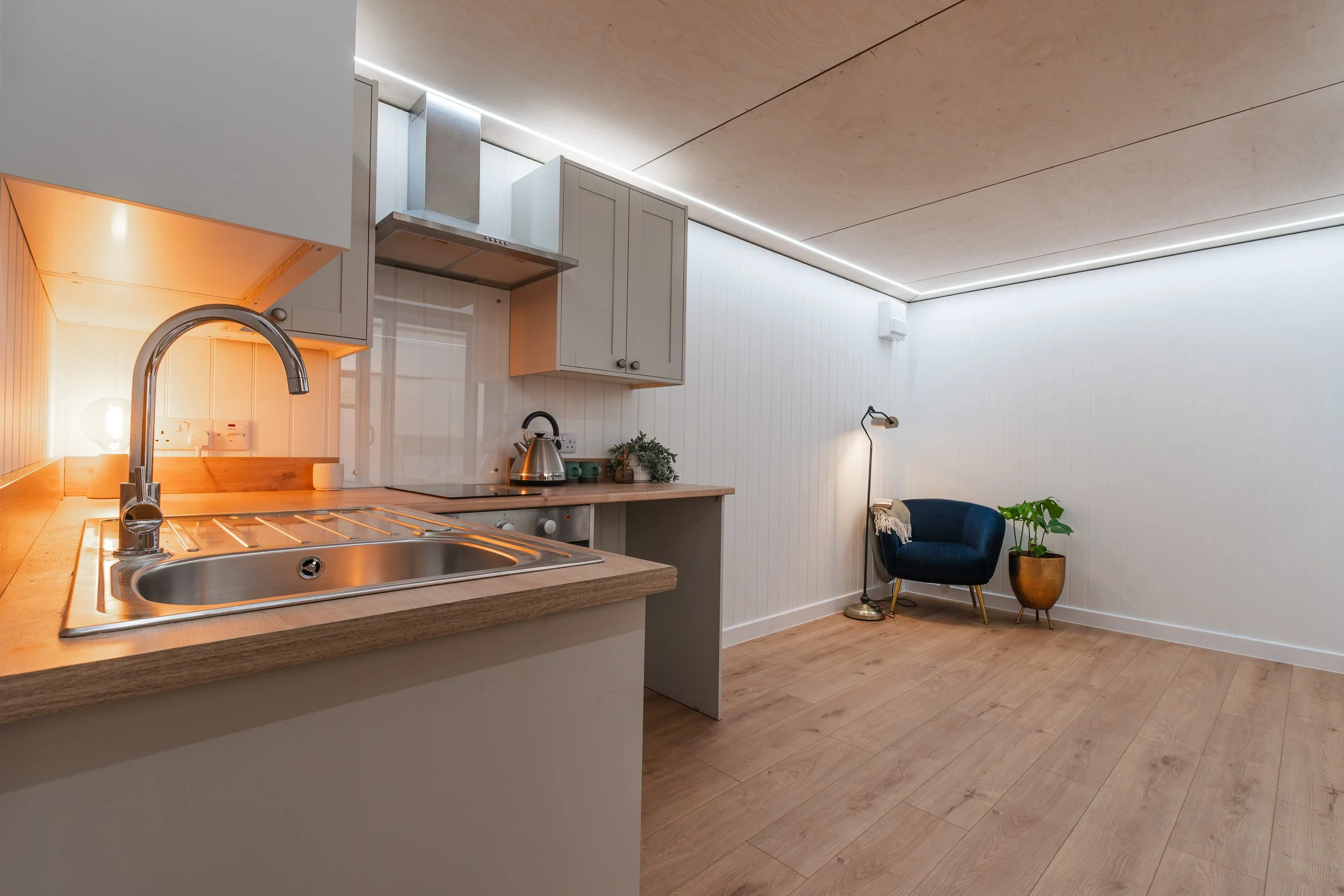 Modern kitchen with light wood flooring, white cabinetry, stainless steel range hood, and a small sitting area with a dark blue chair, a floor lamp, and a potted plant.