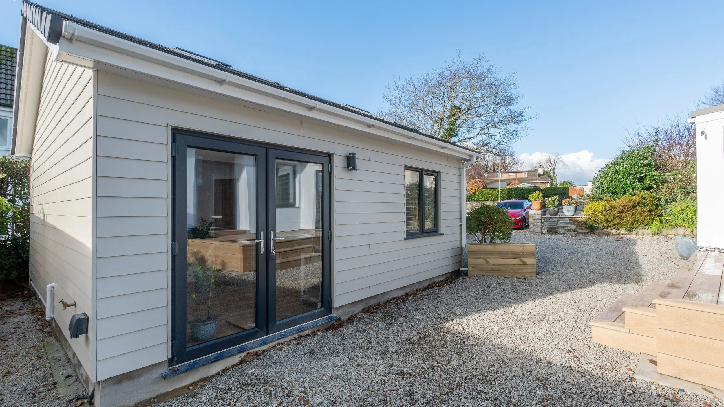 Newly constructed small house with white siding, black framed glass doors, and two black framed windows on gravel yard, with a stepping wooden staircase on the right and a garden with shrubs and pots in the background.