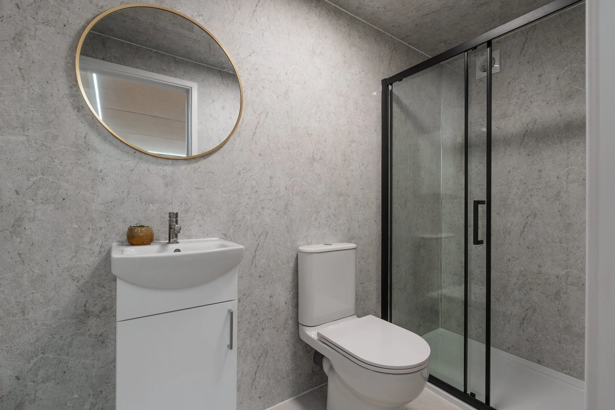 Modern bathroom with a round mirror above a white sink, a ceramic planter, a toilet, and a glass shower enclosure with black framing, all set against gray textured wall tiles.