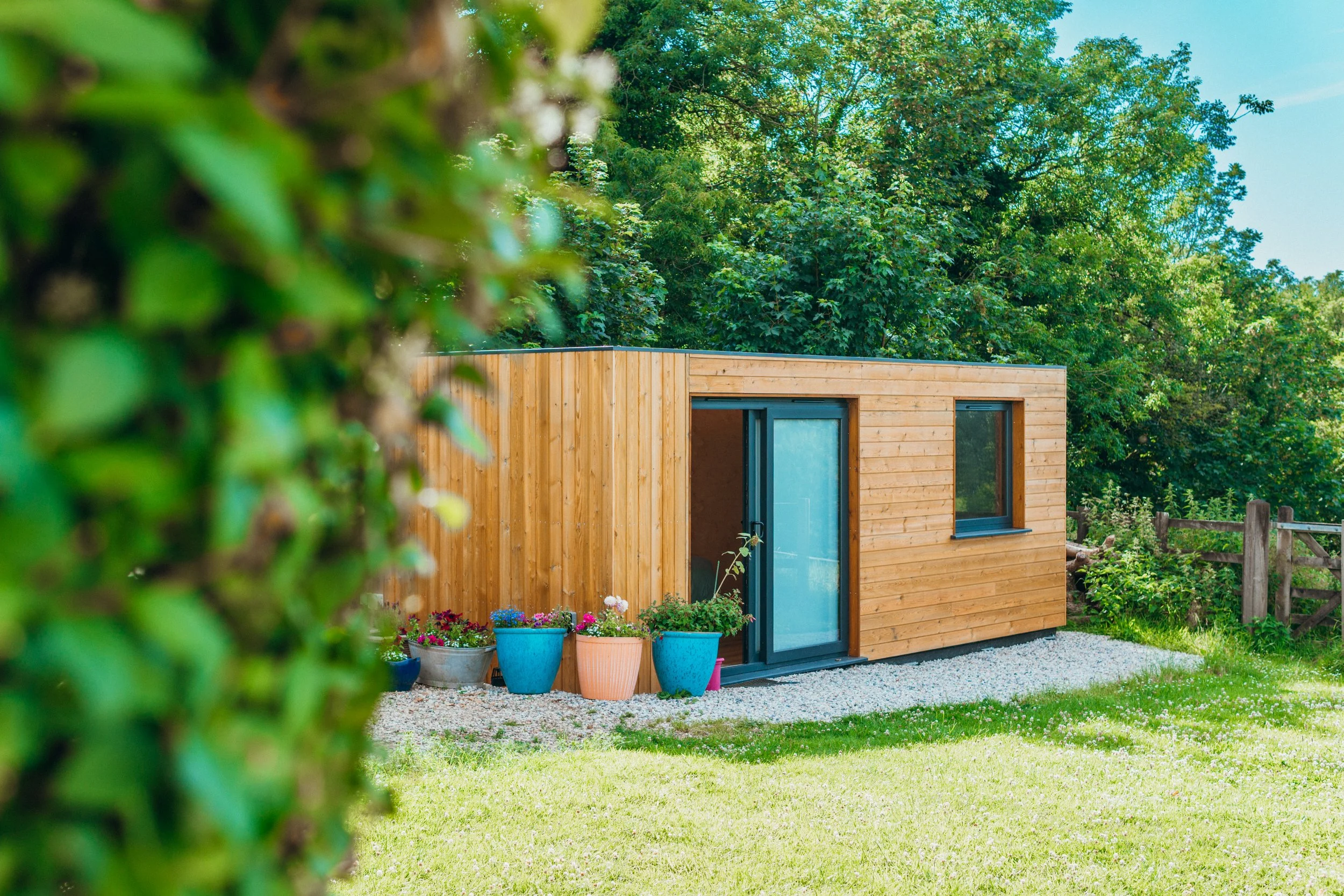 A small wooden house with large windows, surrounded by colorful flower pots, situated in a green garden with trees in the background. The photo is taken through a bush or shrub, partially obscuring the view.