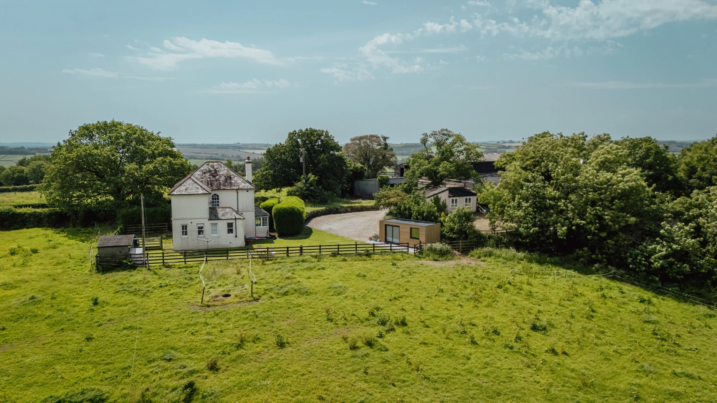 A countryside scene featuring a white house with a slate roof, surrounded by green trees, a fenced grassy yard, a phase garden room, and rolling green fields extending into the distance under a partly cloudy sky.