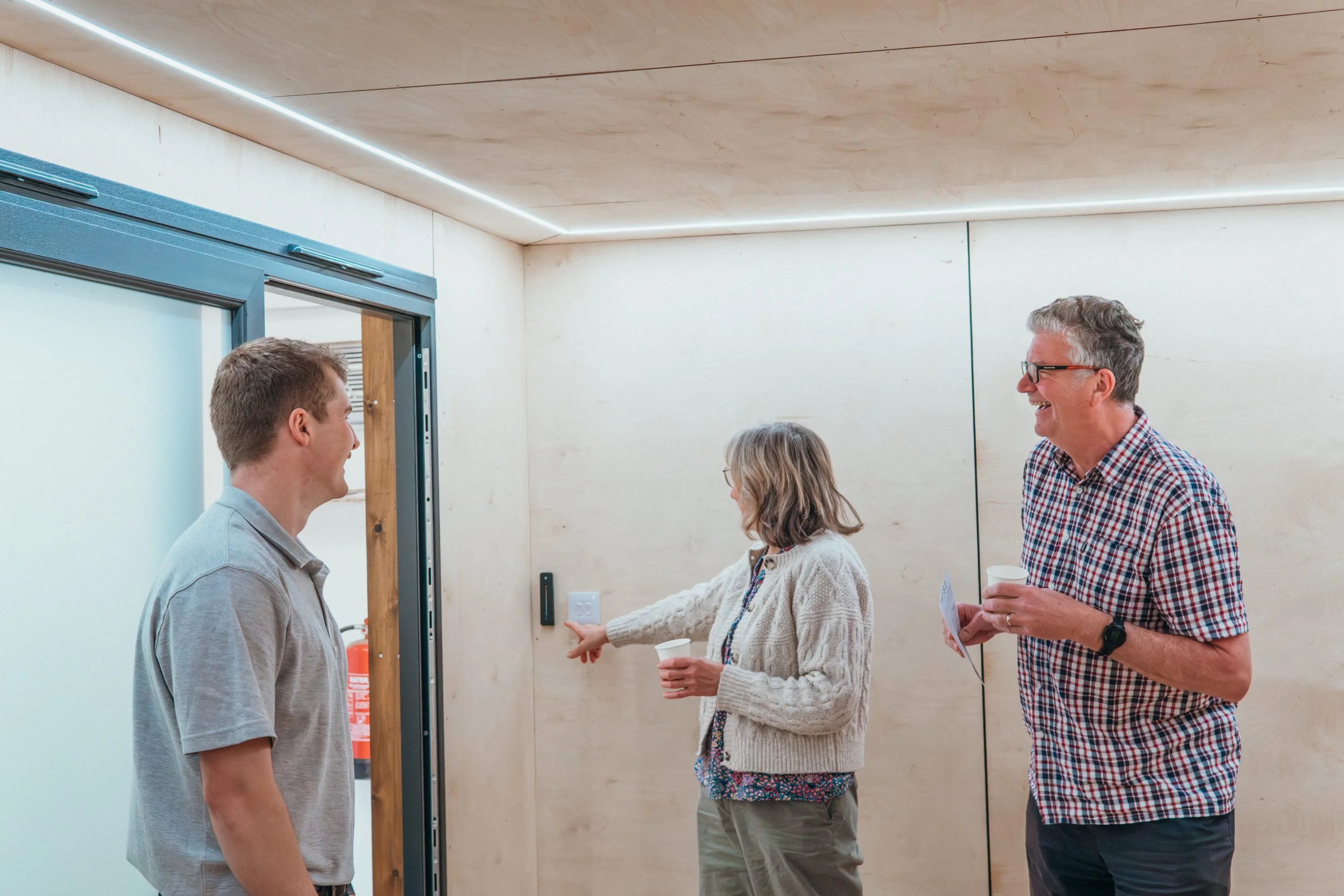 Four people are having a conversation interior garden roof, with glass doors behind them and a plain wall. Two women and two men, some holding paper cups, are smiling and engaging with each other.