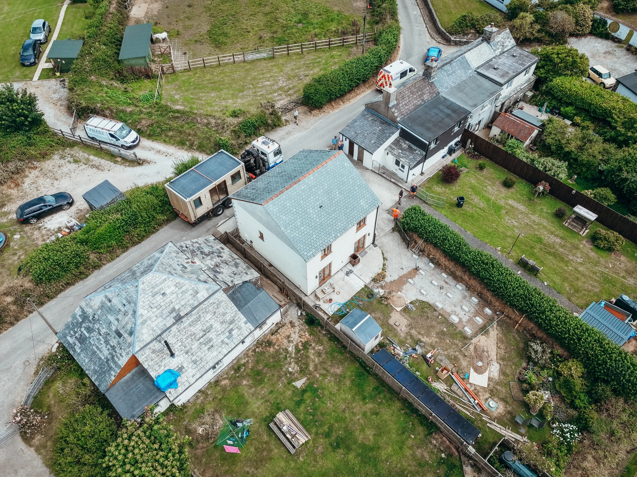 Aerial view of a residential backyard showing a house with a gray roof, a small shed, a fenced garden area with horses, a driveway with parked cars, and a person standing near the house.