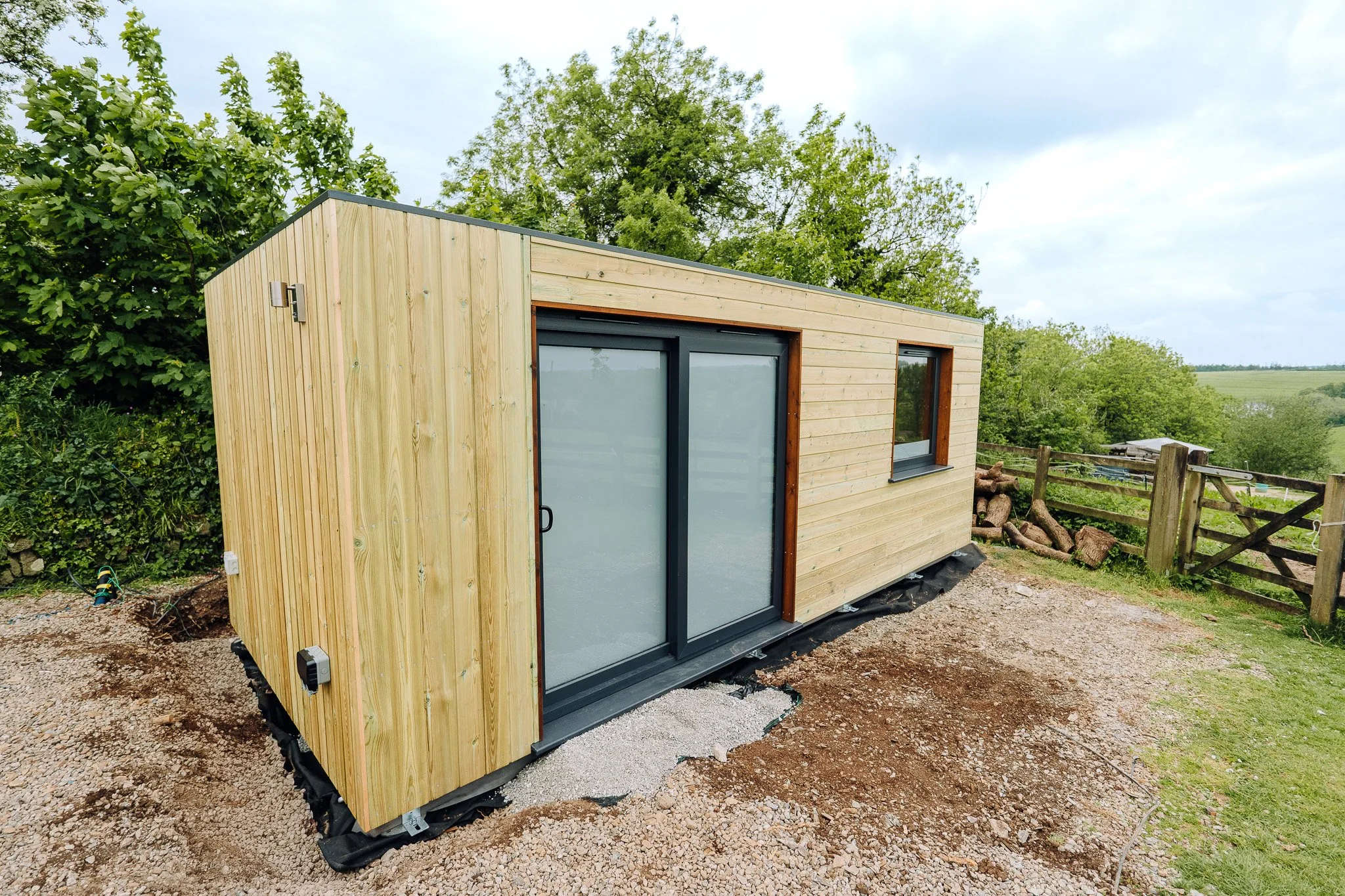 A small wooden tiny house with large sliding glass doors and a window, situated outdoors amidst trees and rural landscape.