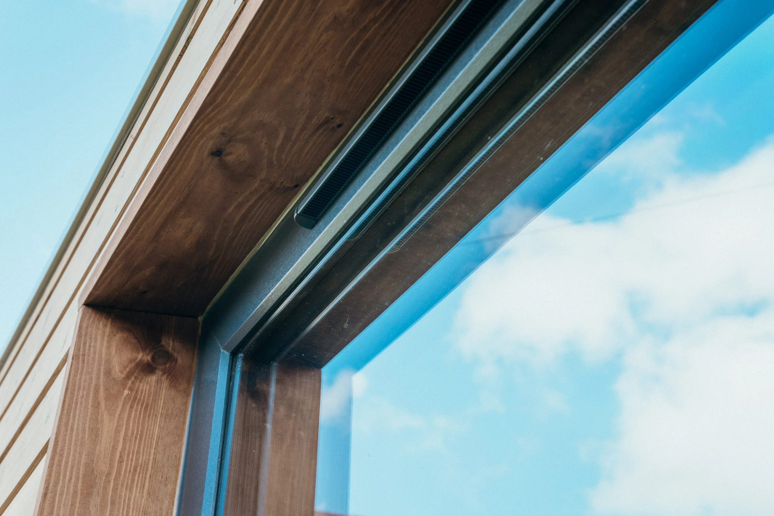 Close-up of a window with a wooden frame and metal vent, showing part of a blue sky with clouds.
