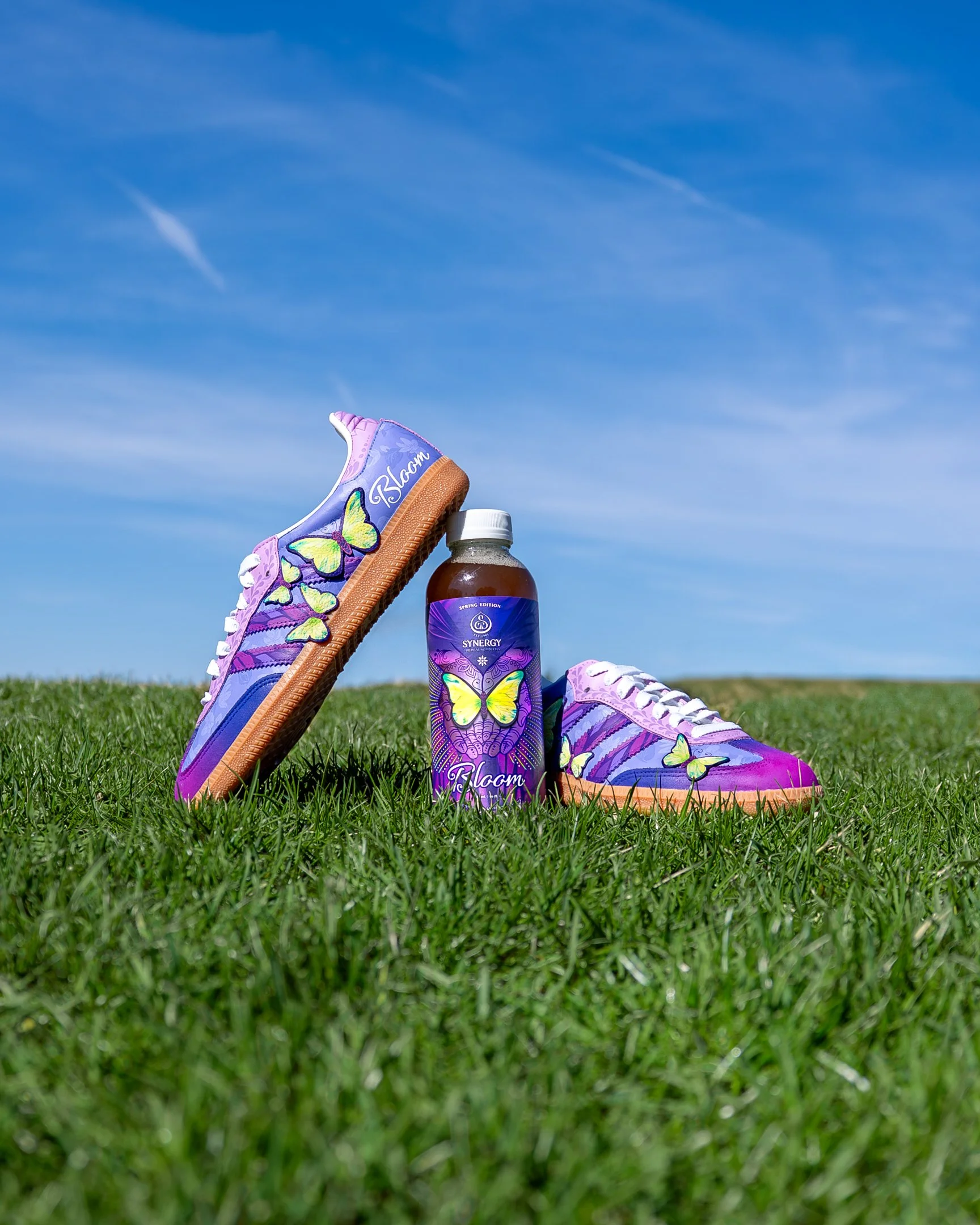 Sneaker Queens Customs Synergy Kombucha “Bloom” custom sneakers and bottle photographed on green grass.