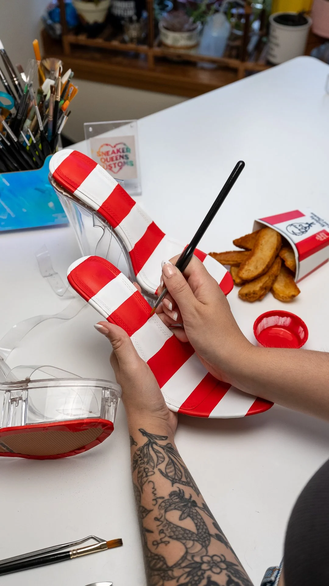 Artist painting red and white KFC bucket stripes on custom high heels beside potato wedges, product photography for KFC x Sneaker Queens Customs.