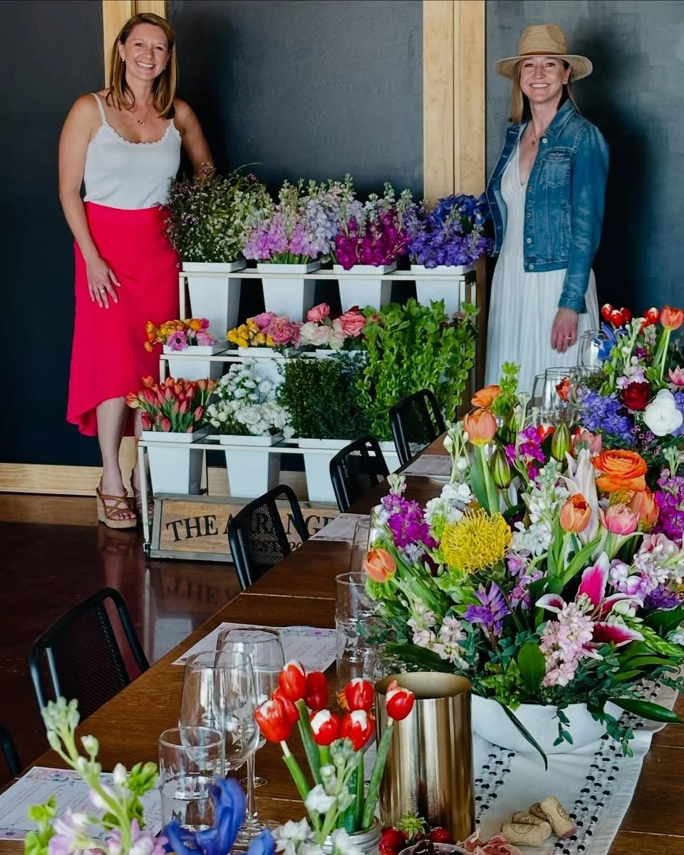 Two women standing behind a display of colorful flowers at a floral event, with a table set with more flower arrangements and empty glasses in the foreground.