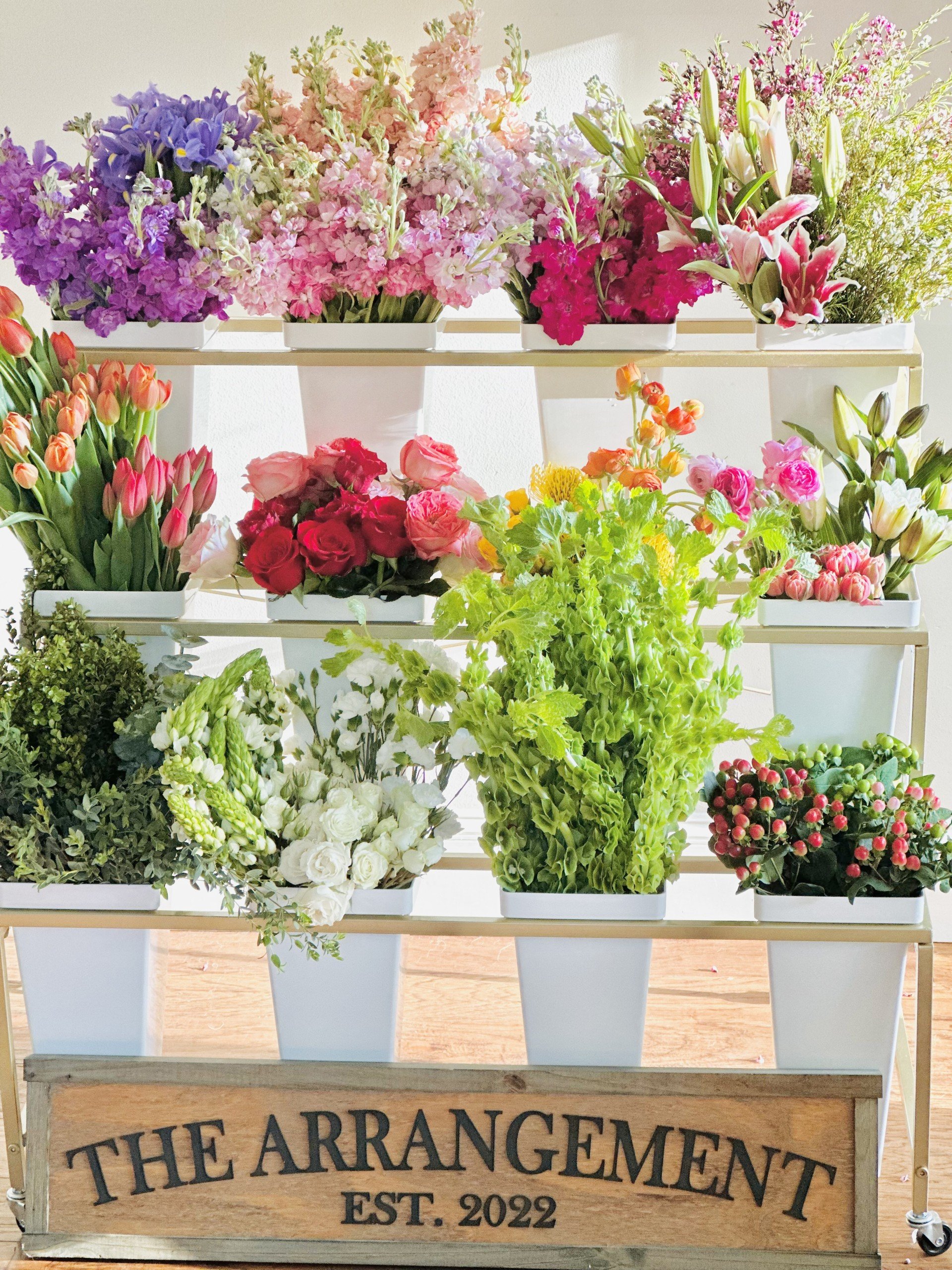 Display of various colorful fresh flowers arranged in white pots on a multi-tiered stand with a wooden sign reading 'The Arrangement Est. 2022' in front.