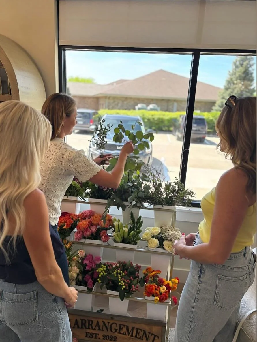Three women arranging flowers at an indoor flower shop, with a large window showing parked cars and a building outside.
